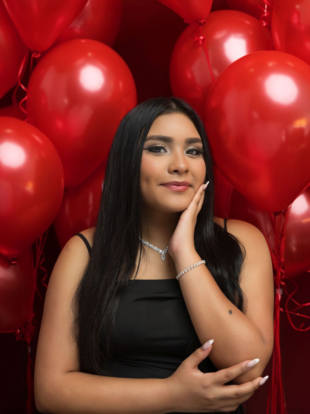 A glamour portrait against red balloons wearing a black top and sparkly jewelry.