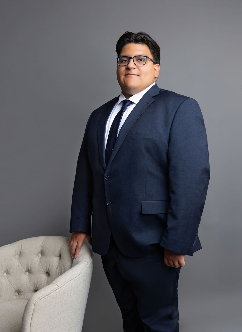 Business headshot of person in dark blue suit posing next to grey tufted chair.