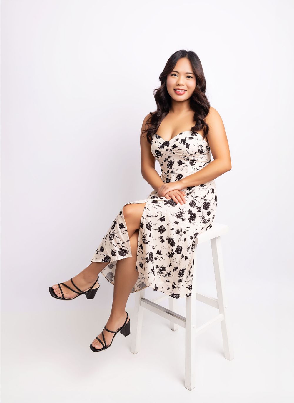 Person in floral dress poses elegantly on white stool against white background.