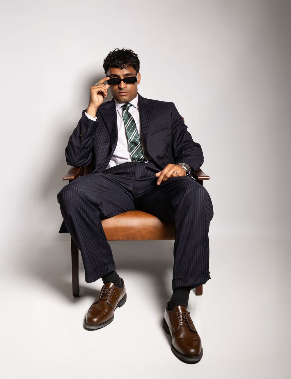 Professional in pinstriped suit and sunglasses sits confidently in leather chair wearing green striped tie and brown shoes.
