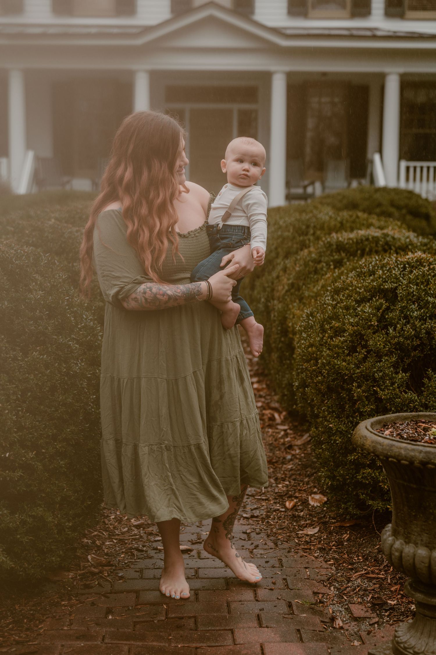 A woman in a flowing olive dress stands barefoot on brick pavers while holding a baby outside a house. photo by Samantha Campbell imagery