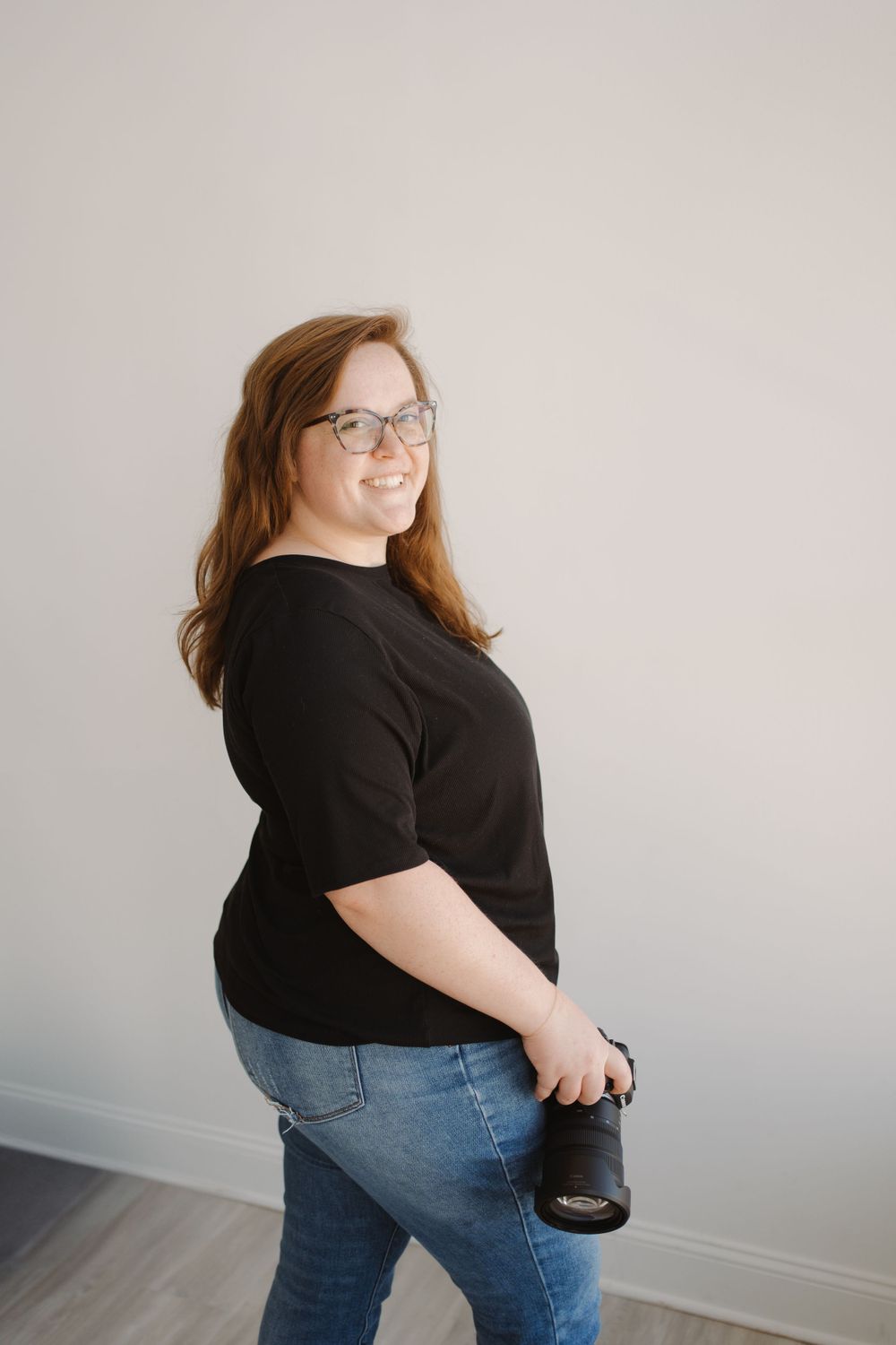 A person wearing glasses in a black top and jeans holding a camera while posing against a gray wall.