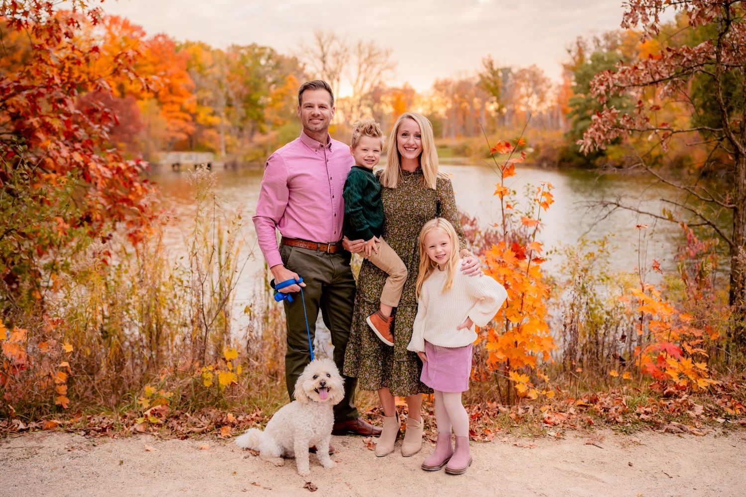 A family and their white dog pose together by a lake surrounded by autumn foliage.