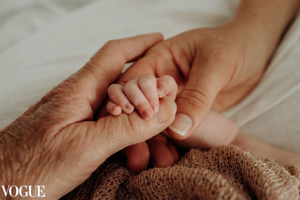 A tiny baby hand grasps an adult finger while wrapped in a brown blanket with Vogue photography watermark.