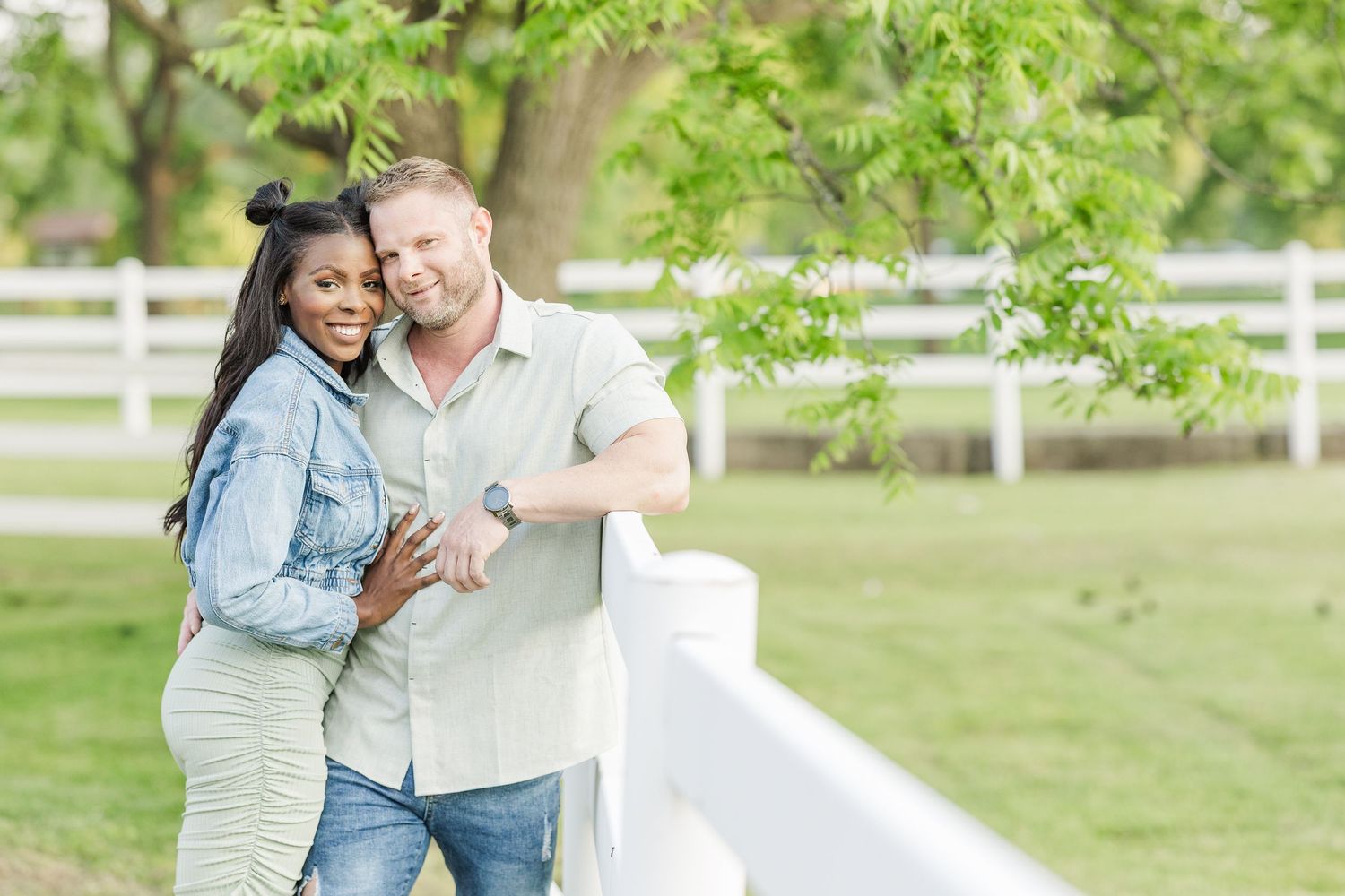 Happy couple embraces by white fence in sunny garden with green trees and grass.