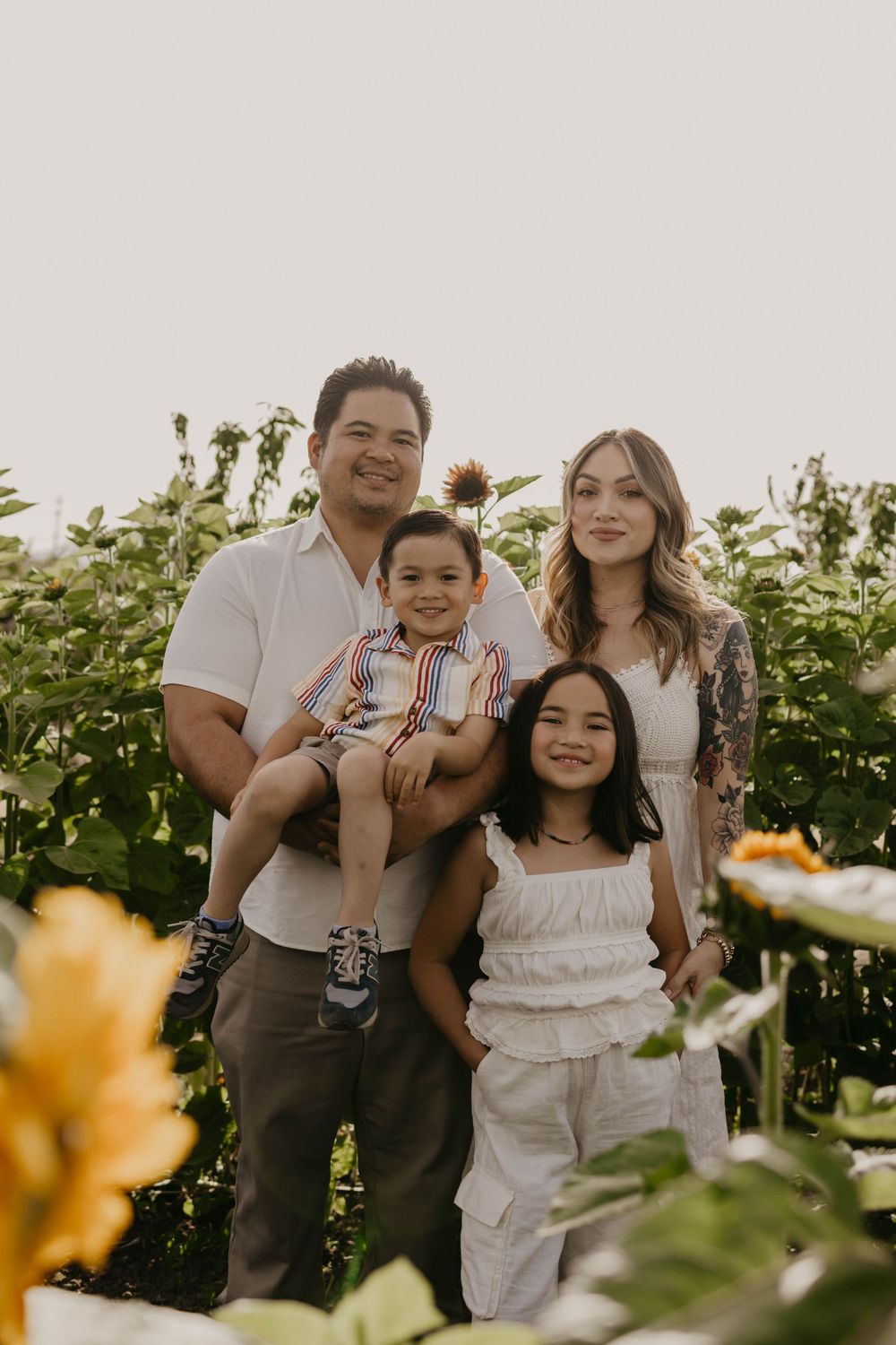 A family poses together in a garden with sunflowers and greenery on a bright summer day.