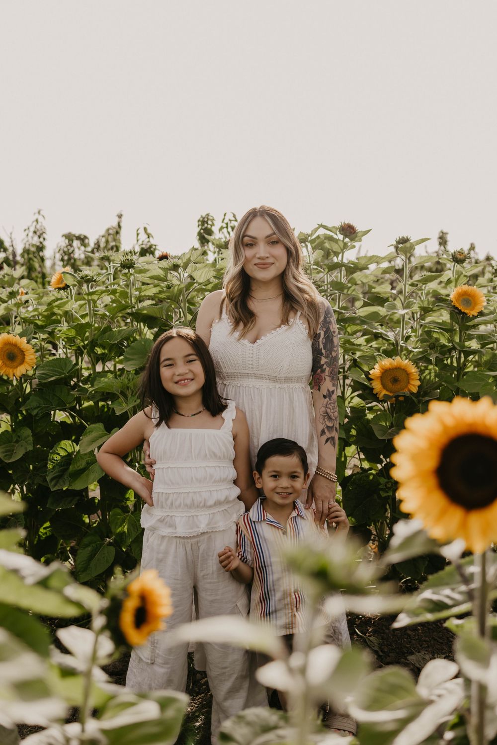 Four people in white and neutral colored outfits pose together in a blooming sunflower field during sunset.