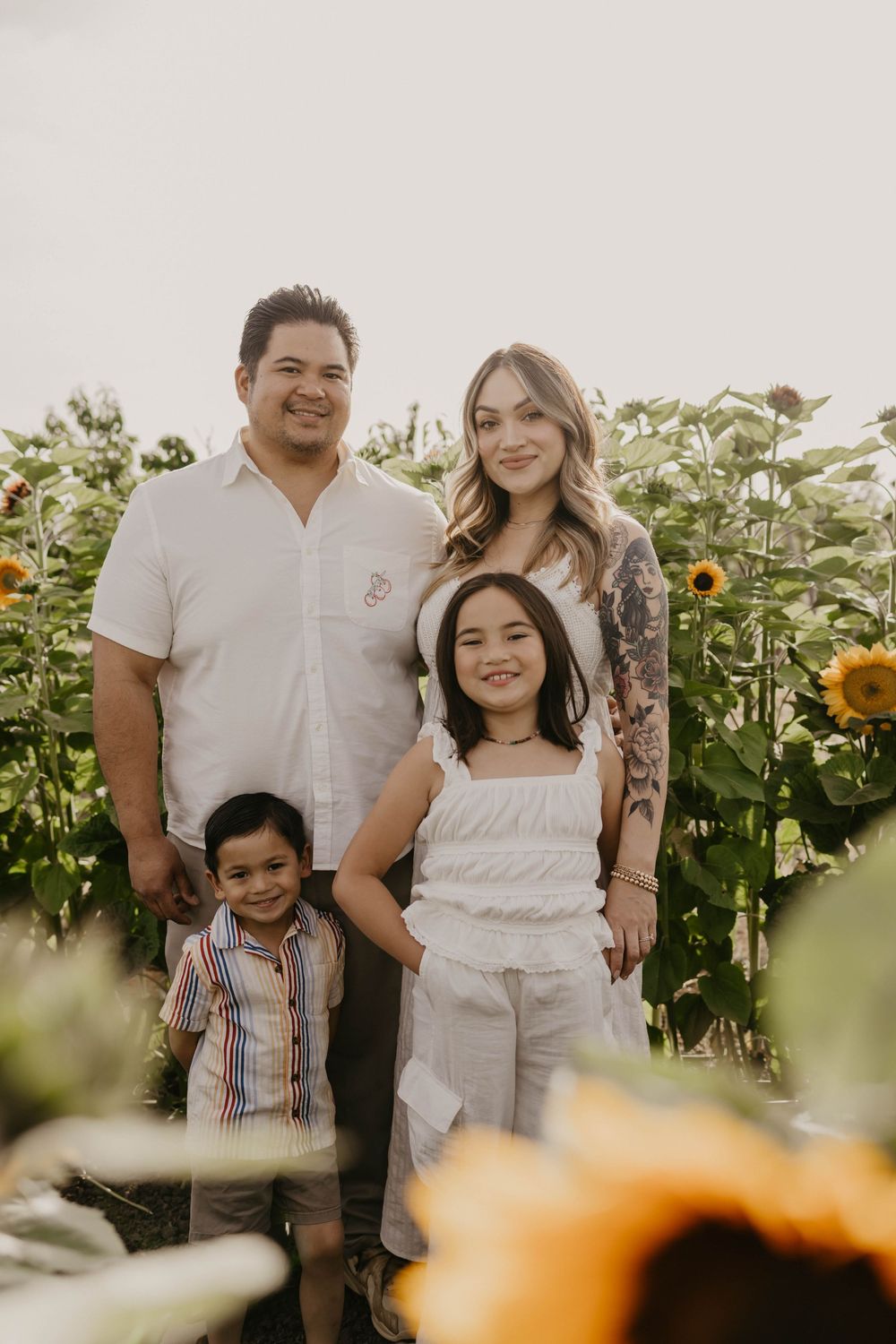 A family poses together in a sunflower field on a bright sunny day, surrounded by tall blooming sunflowers.