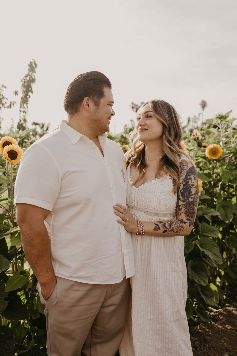 A couple shares an intimate moment in a sunflower field at golden hour wearing white and neutral colored outfits.