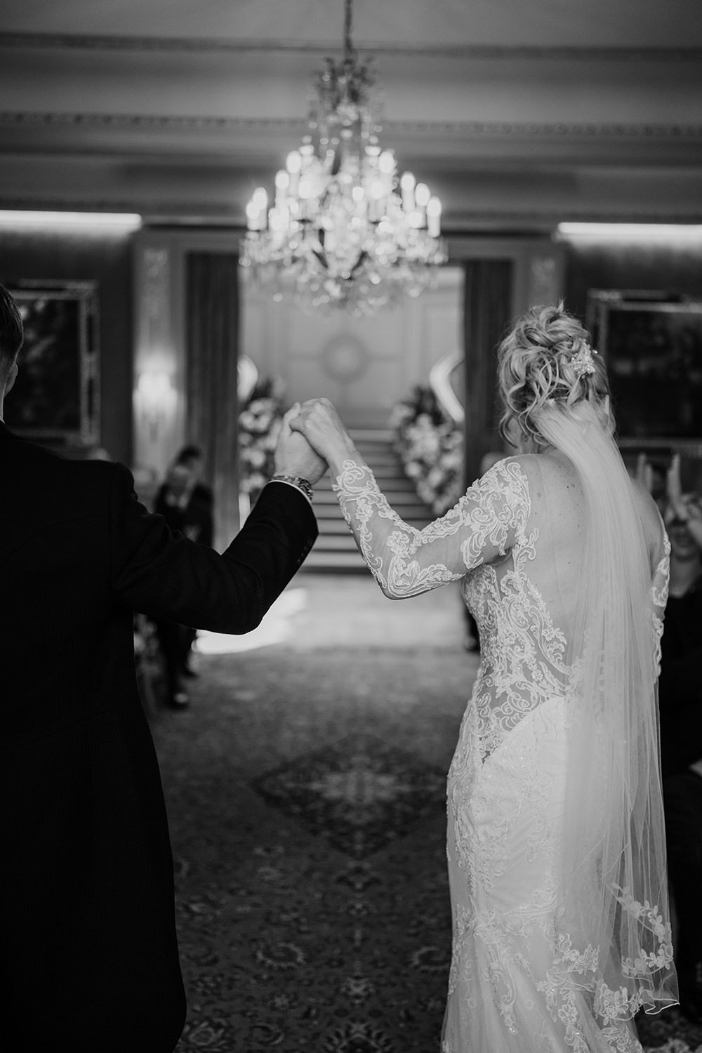 A bride in a lacy wedding dress and veil dances with her partner under an elegant crystal chandelier in a formal ballroom.