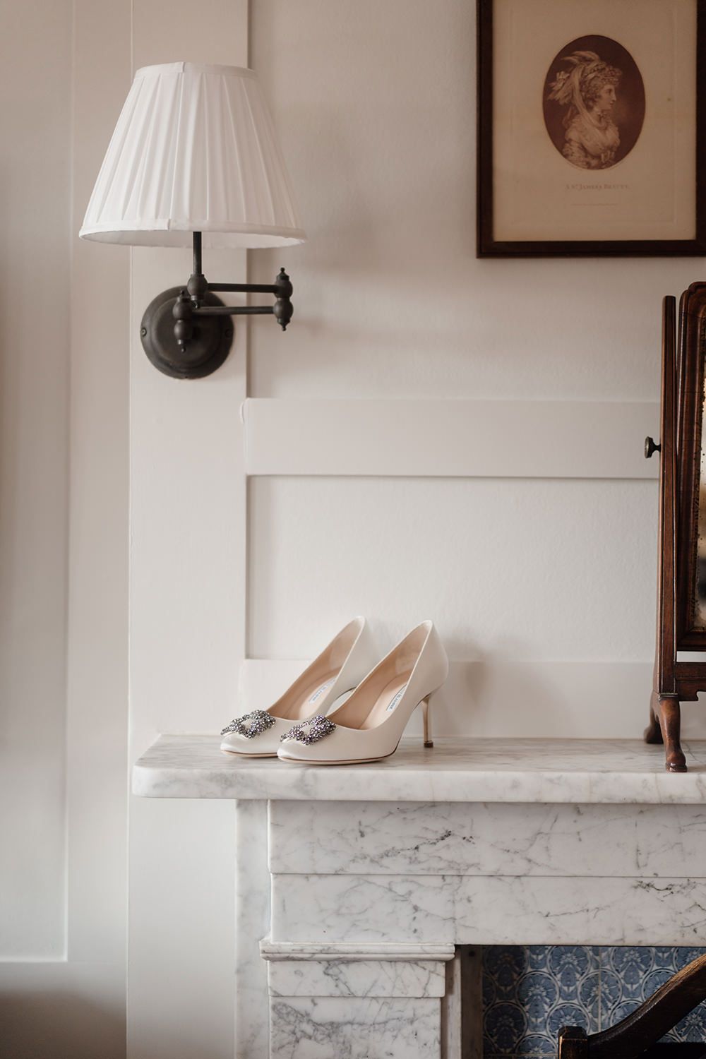 A pair of elegant white bridal shoes displayed on a marble mantelpiece with table lamps and framed artwork.