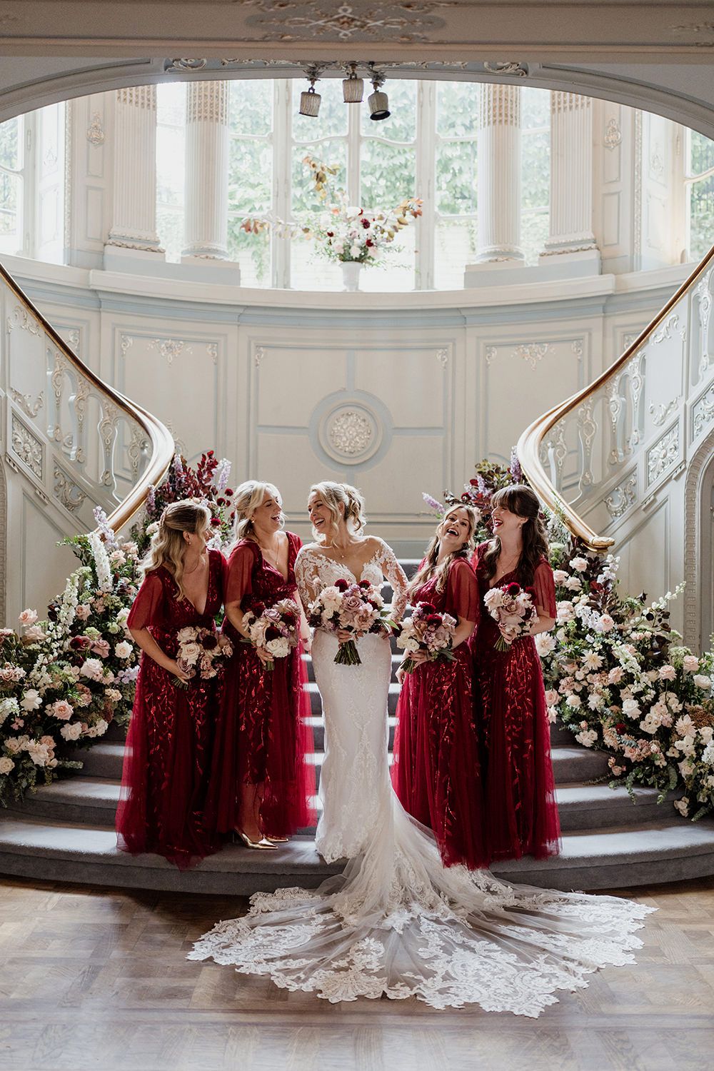 Wedding party in burgundy dresses poses against white columns with floral arrangements at elegant venue.