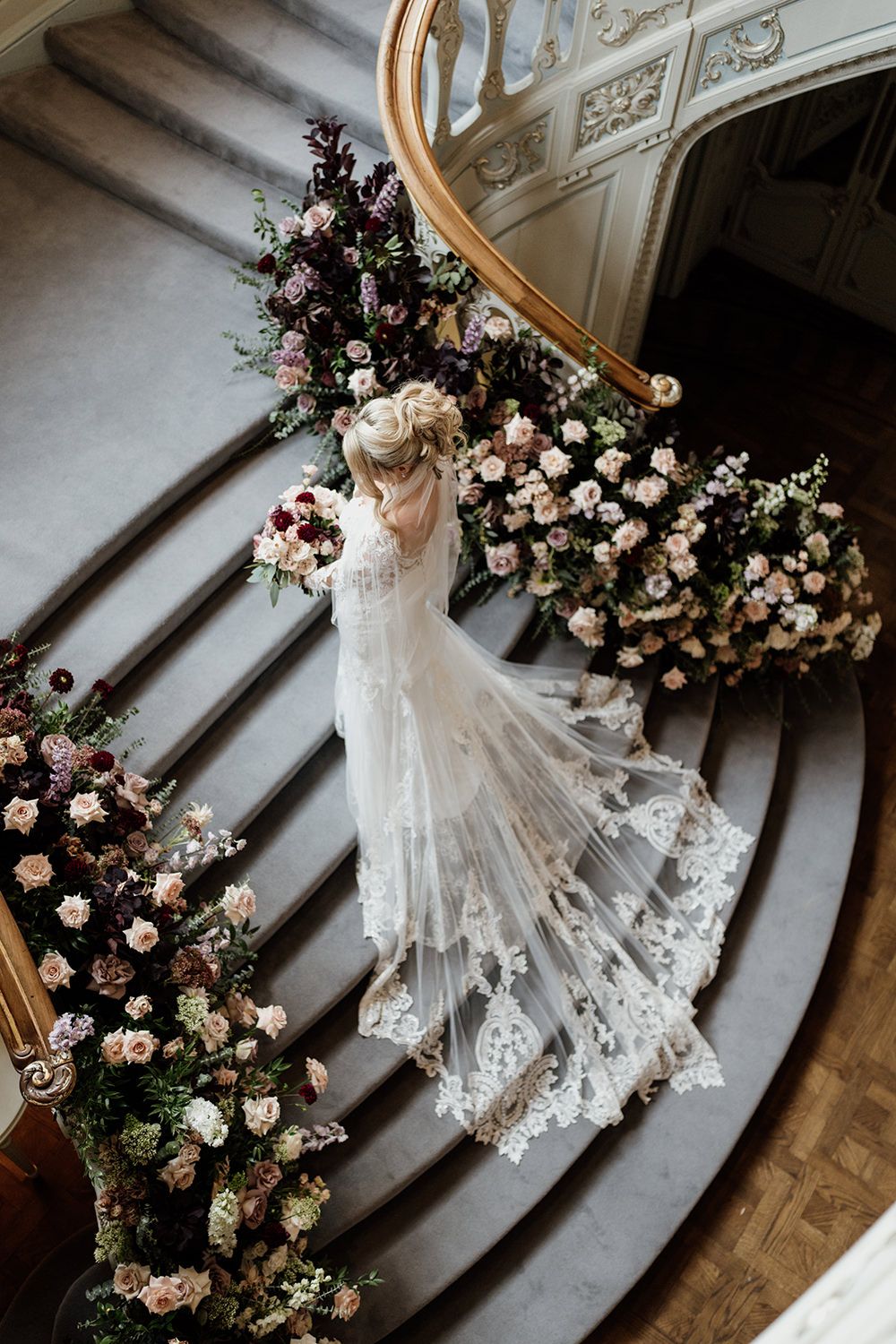 Elegant wedding dress train with delicate lace details trails down ornate staircase near floral arrangements.