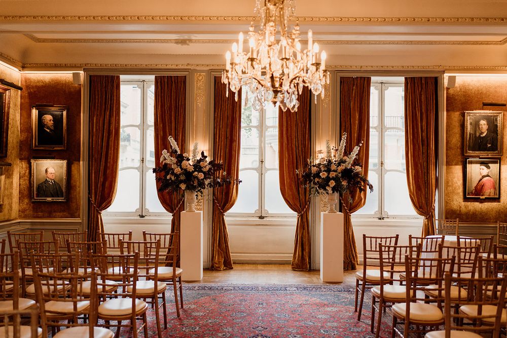 Elegant wedding ceremony setup in grand room with crystal chandelier, wooden chiavari chairs and floral arrangements.