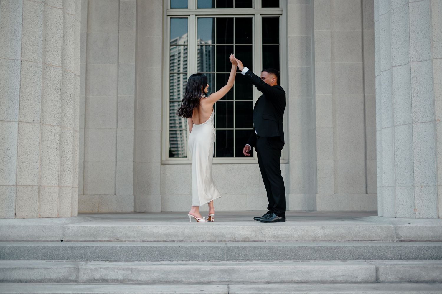 A couple in wedding attire dance together on stone steps between elegant white columns.