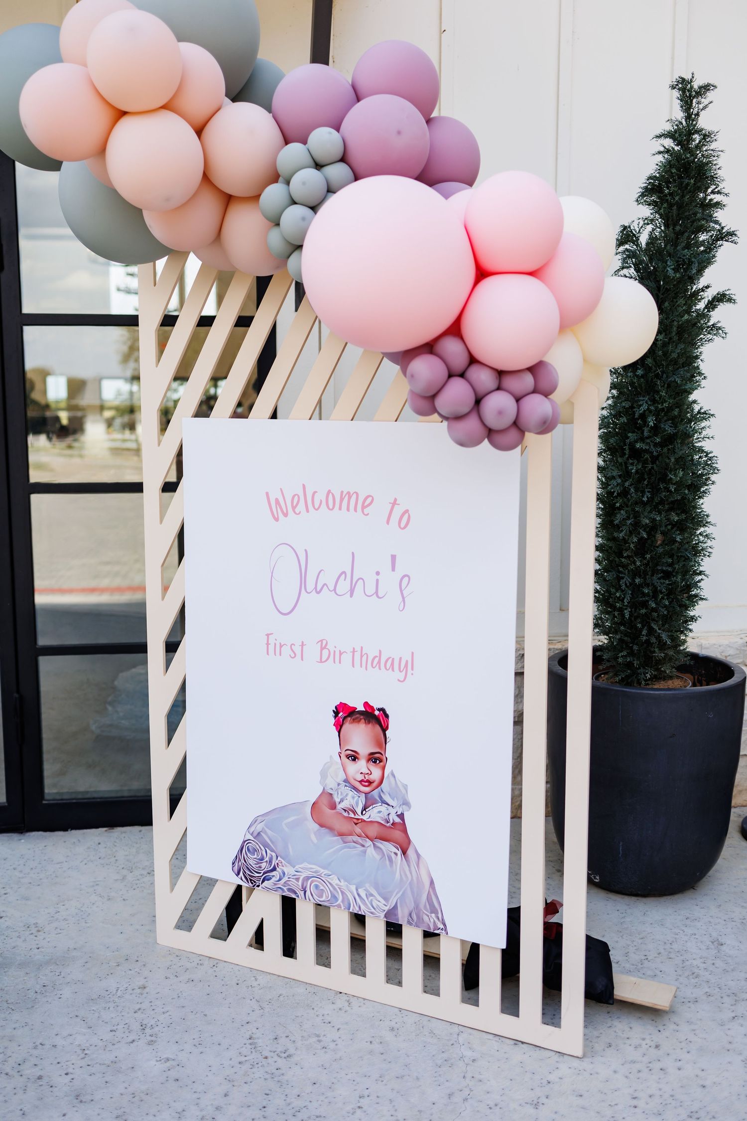 Decorative balloon arch in soft pastel pink, purple, and peach colors with a welcome birthday sign on a white slatted display.