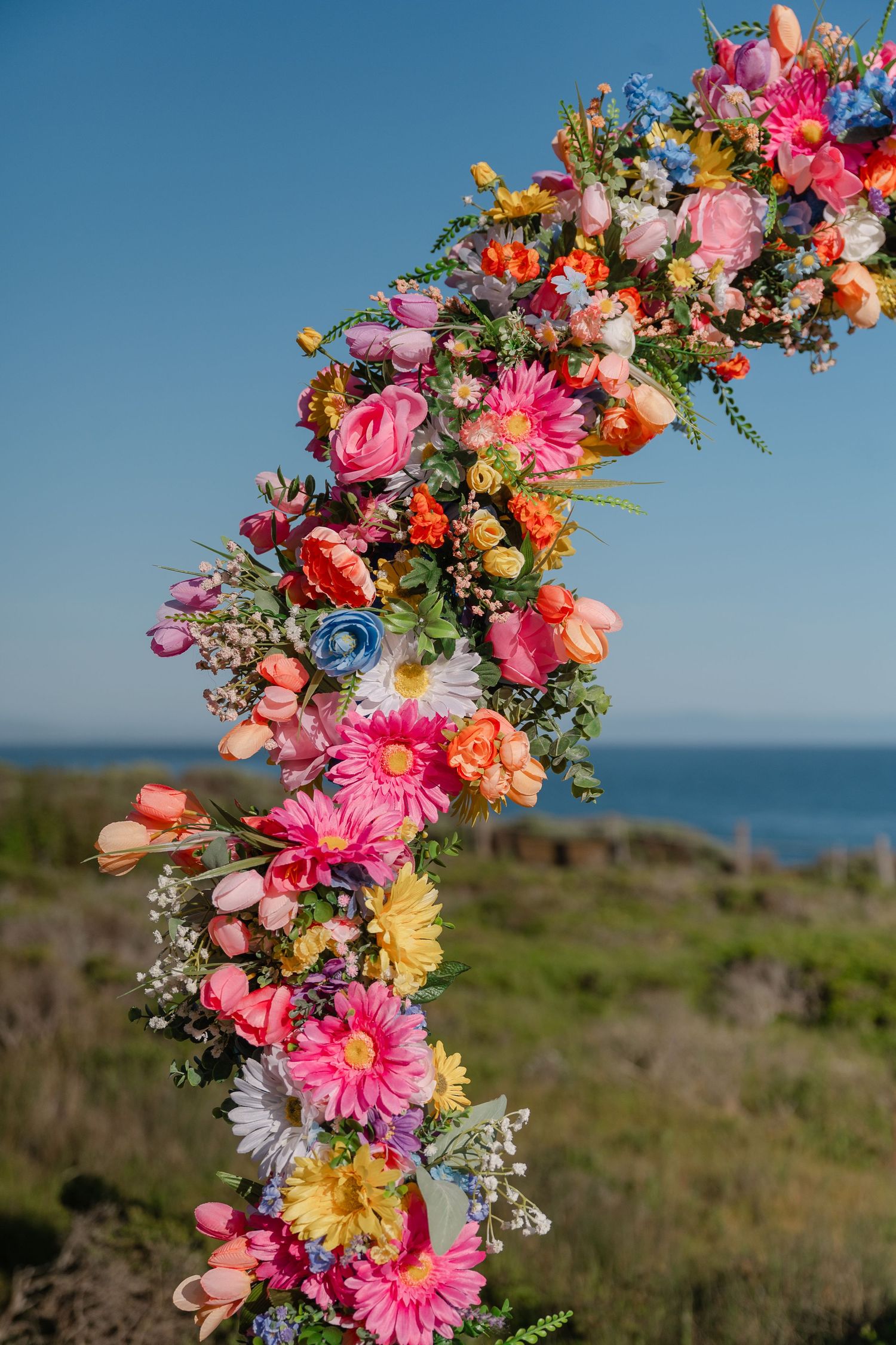 Colorful floral arch installation with pink, orange and yellow blooms against blue ocean and sky backdrop.