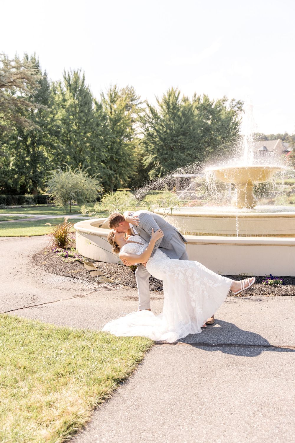 A romantic dip pose being performed by a couple in formal wedding attire near a garden fountain on a sunny day.