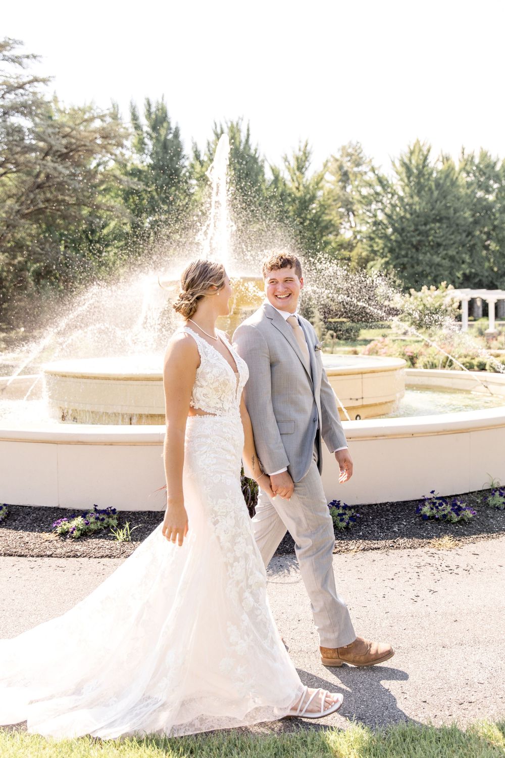 A couple poses in front of a fountain on their wedding day, with sunlit water spray creating a magical backdrop.