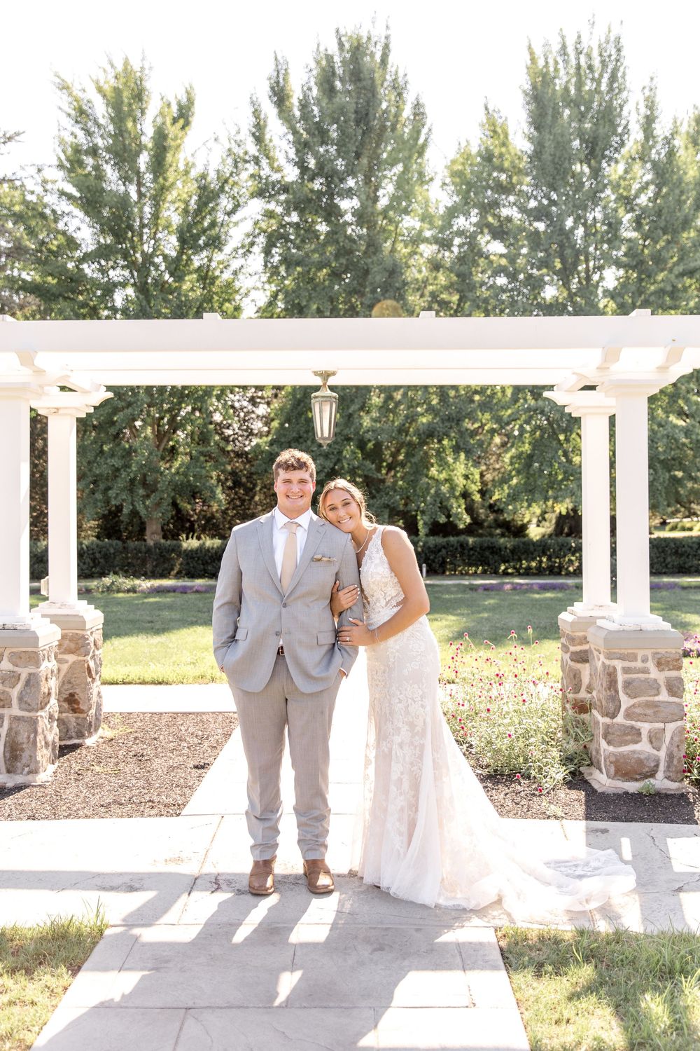 A couple stands under an elegant white pergola with stone pillars on a sunny garden path during their wedding celebration.