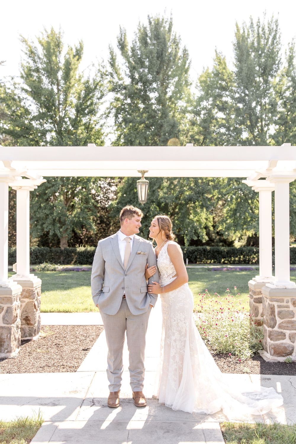 A couple stands under an elegant white pergola with stone pillars in a garden on a sunny summer day.