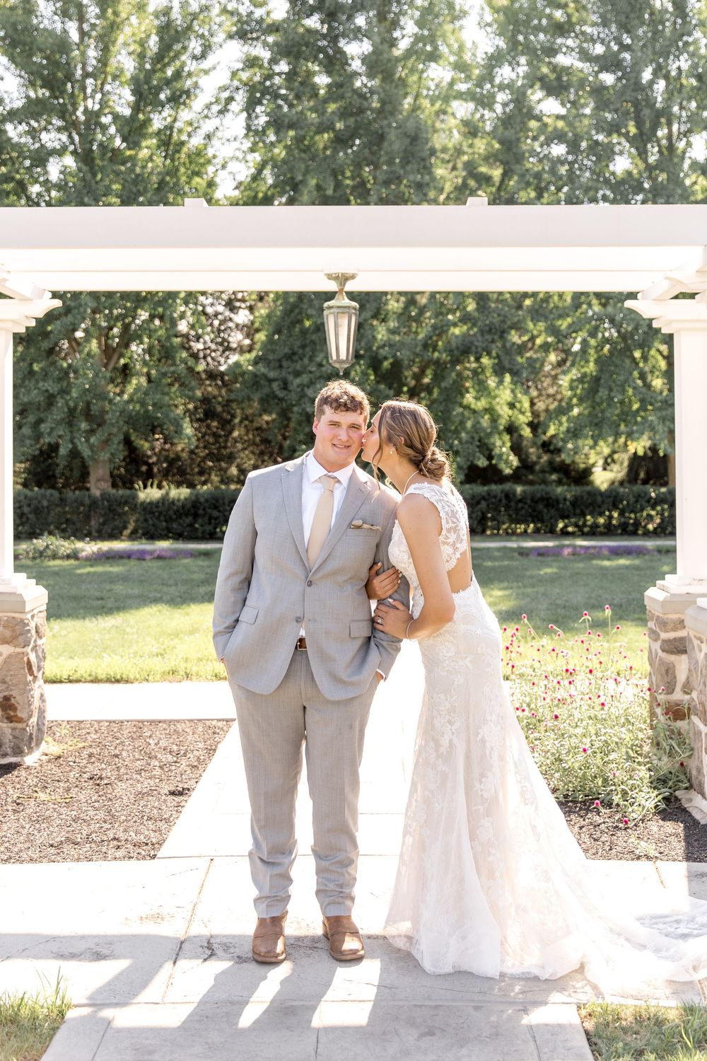 A wedding couple poses under a white pergola on a stone patio during a sunny outdoor ceremony.