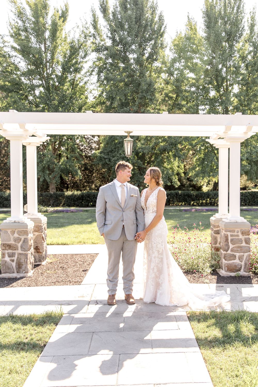 Elegant outdoor wedding scene under a white pergola with stone pillars and lush greenery in the background.