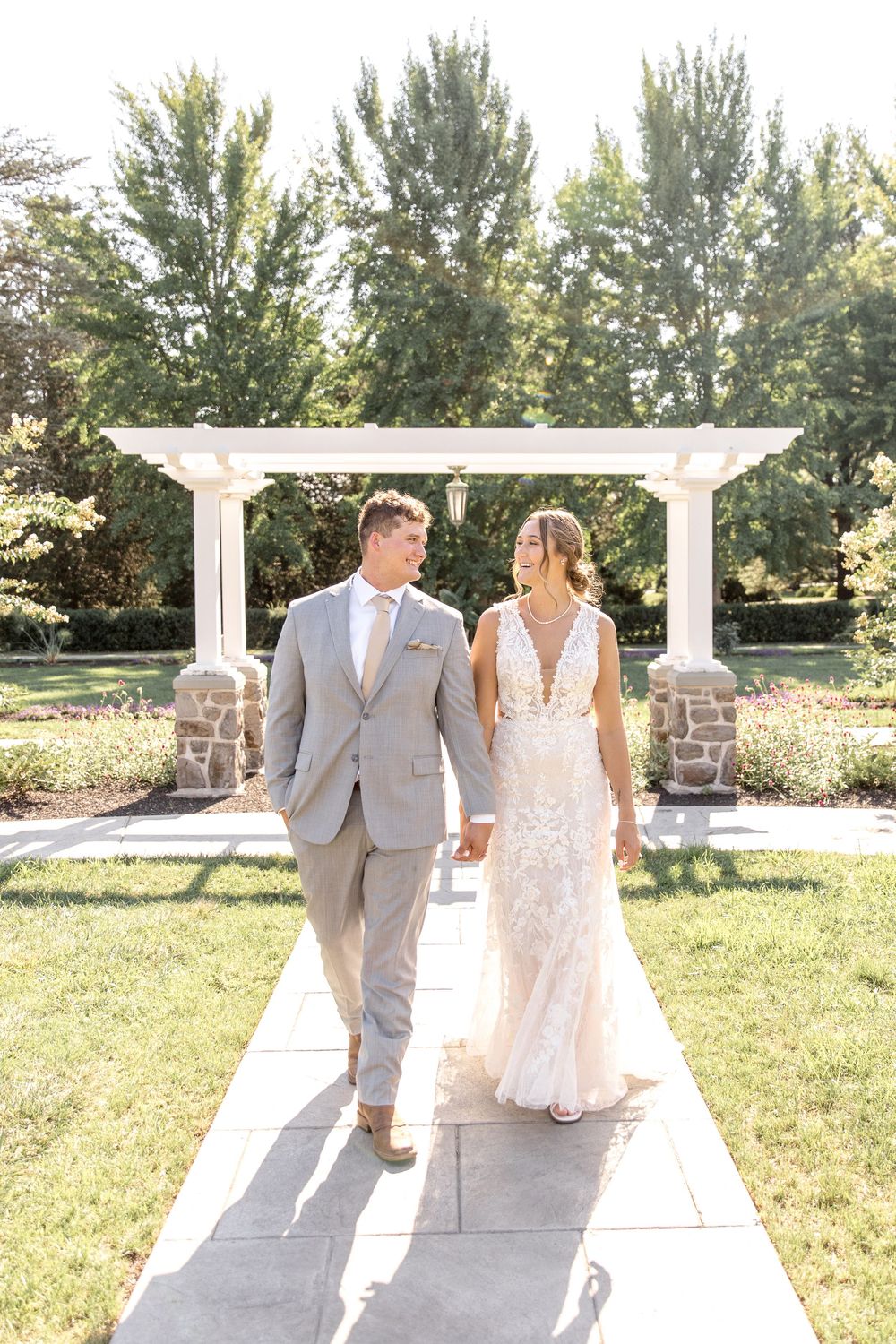 A couple walks along a stone pathway under a white garden pergola on a sunny summer day surrounded by trees.