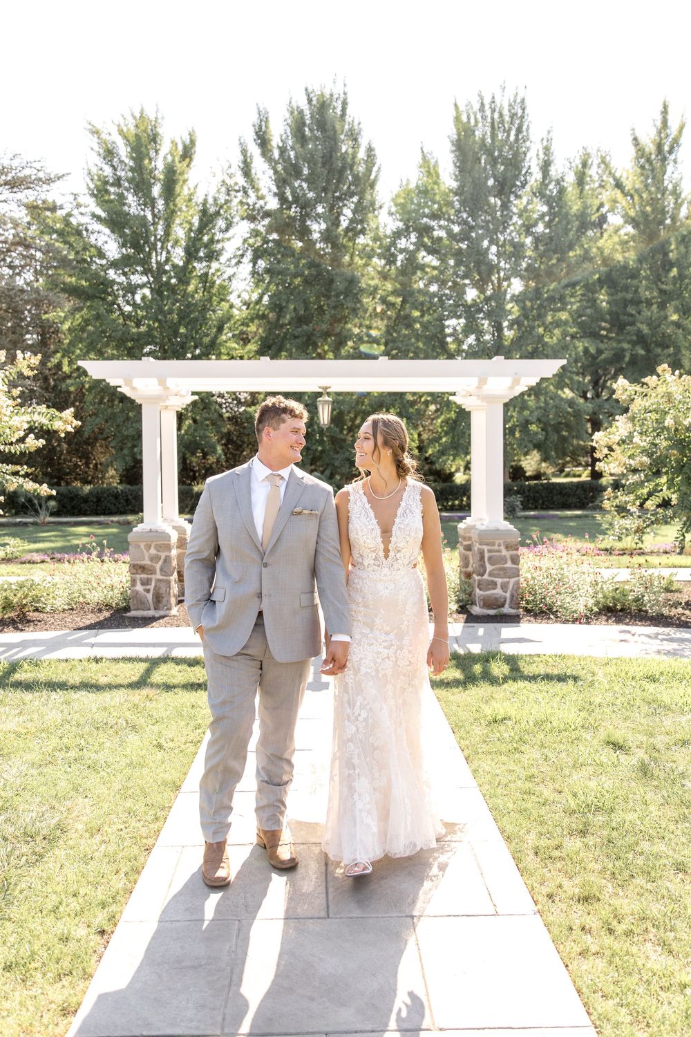 A panoramic view of a wedding couple walking along a sunny outdoor pathway surrounded by green trees and grass.