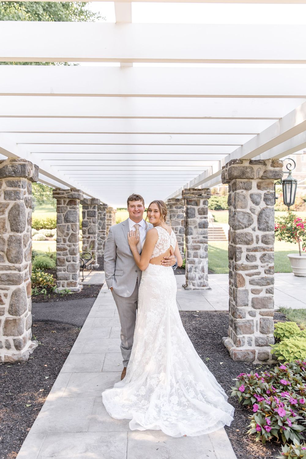 Elegant couple poses under stone pergola with white beams and pink flowers during outdoor wedding ceremony.