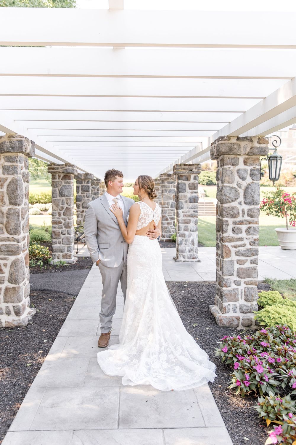 A wedding couple shares a romantic moment under a covered stone walkway with blooming flowers in the garden.