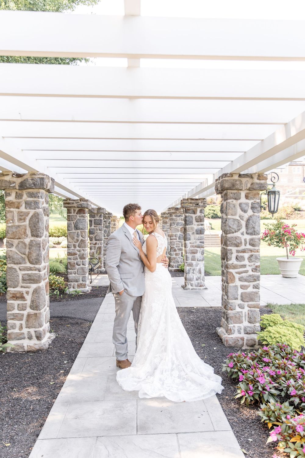Elegant wedding couple dancing together in romantic garden walkway with stone pillars and pink flowers.
