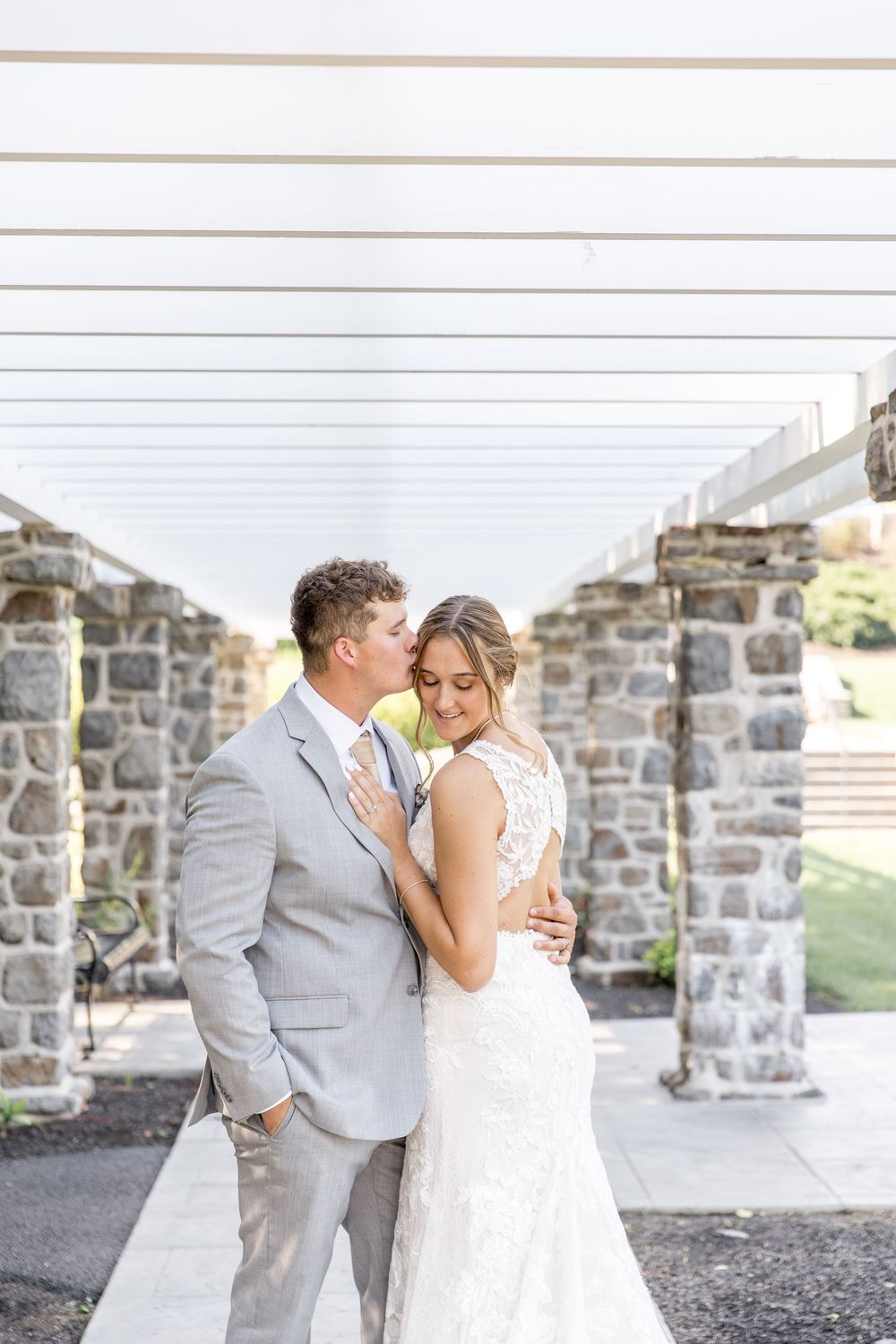 Elegant couple embraces near stone wall archway during romantic outdoor wedding photoshoot.