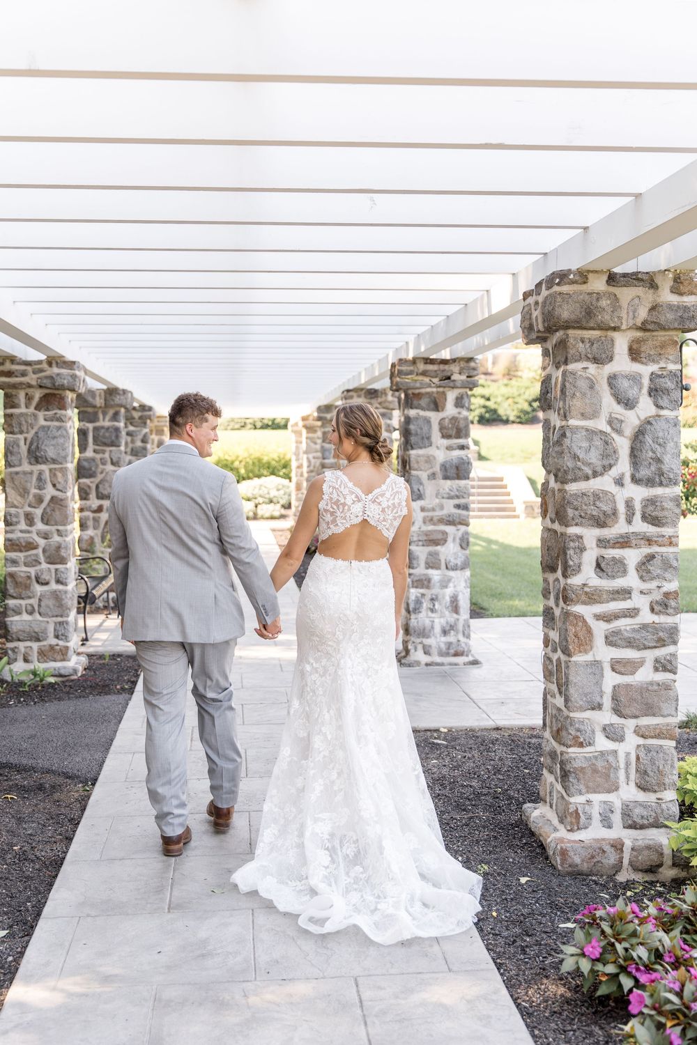 A couple in formal wedding attire walk together under a covered stone pillar walkway with flowers in the garden.