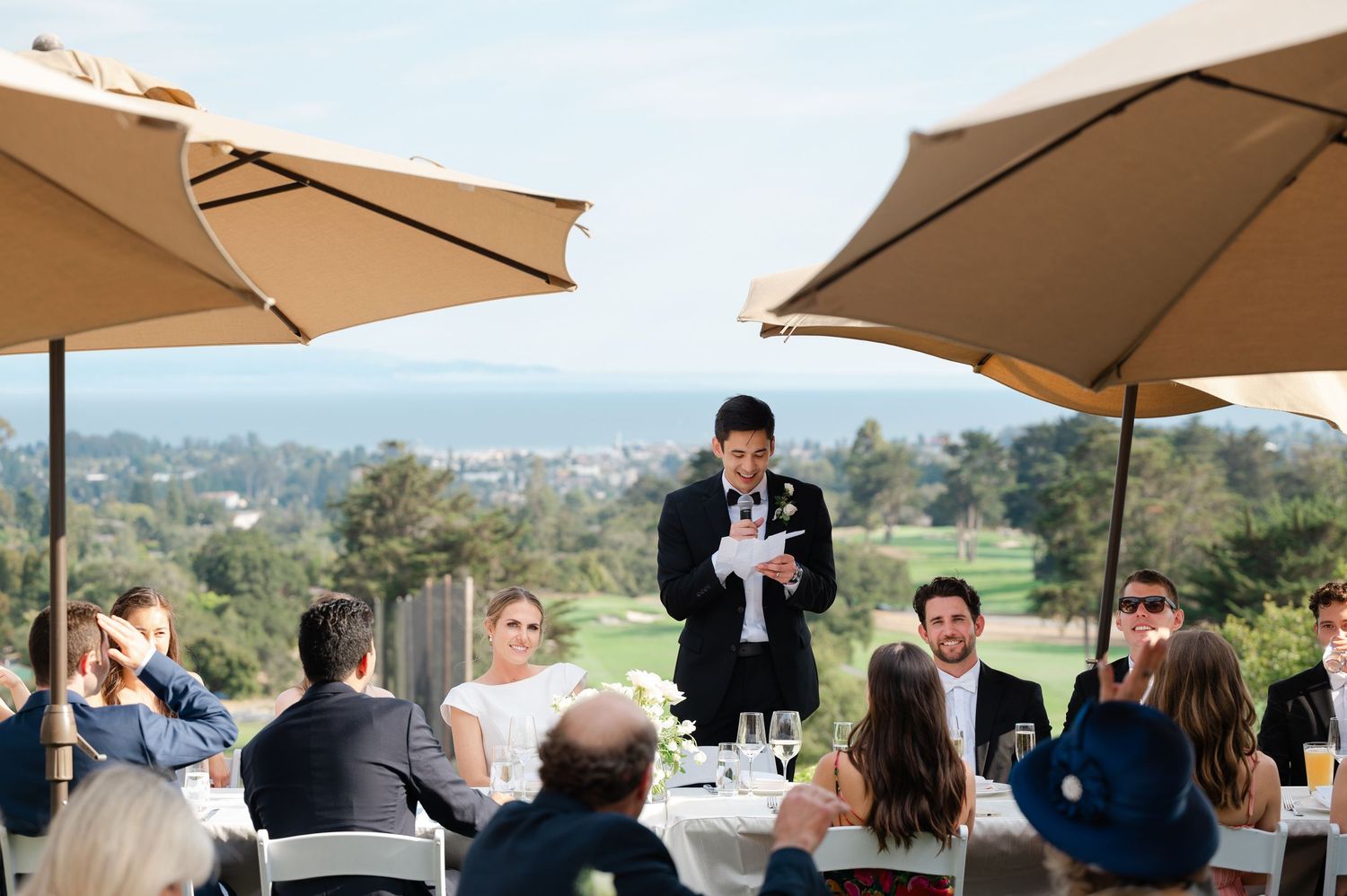 Group gathered for an outdoor event under tan umbrellas on a patio overlooking scenic hills and distant landscape.