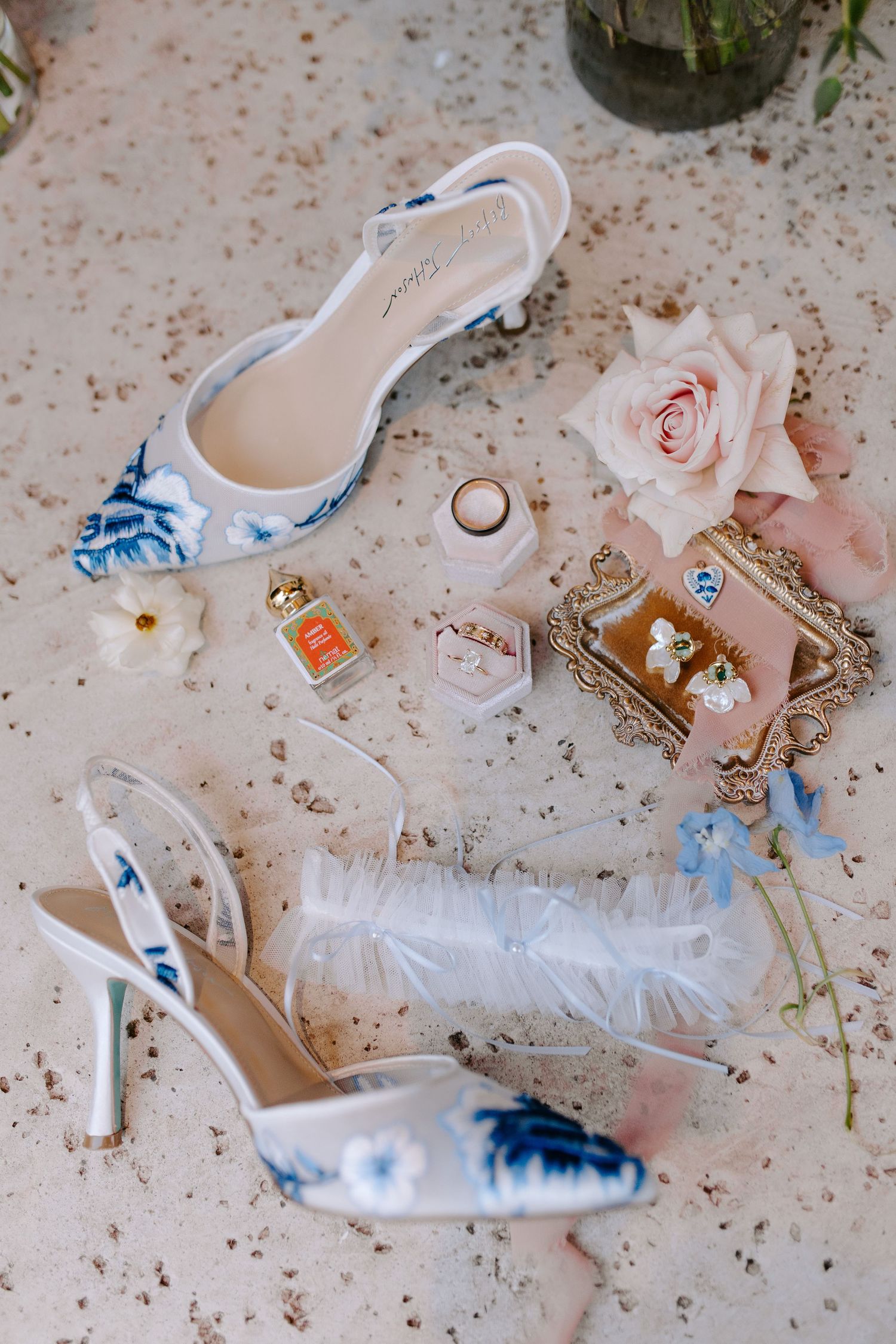 Wedding accessories including blue and white floral heels, rings, perfume, and a delicate pink rose arranged on a textured surface.