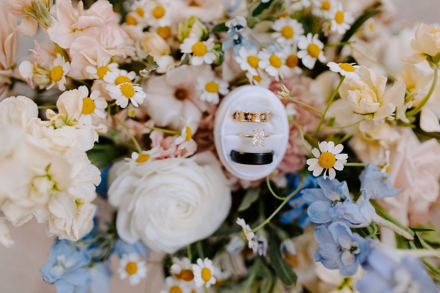 Wedding rings nestled among soft pink roses, white daisies, and blue hydrangeas in an elegant floral arrangement.