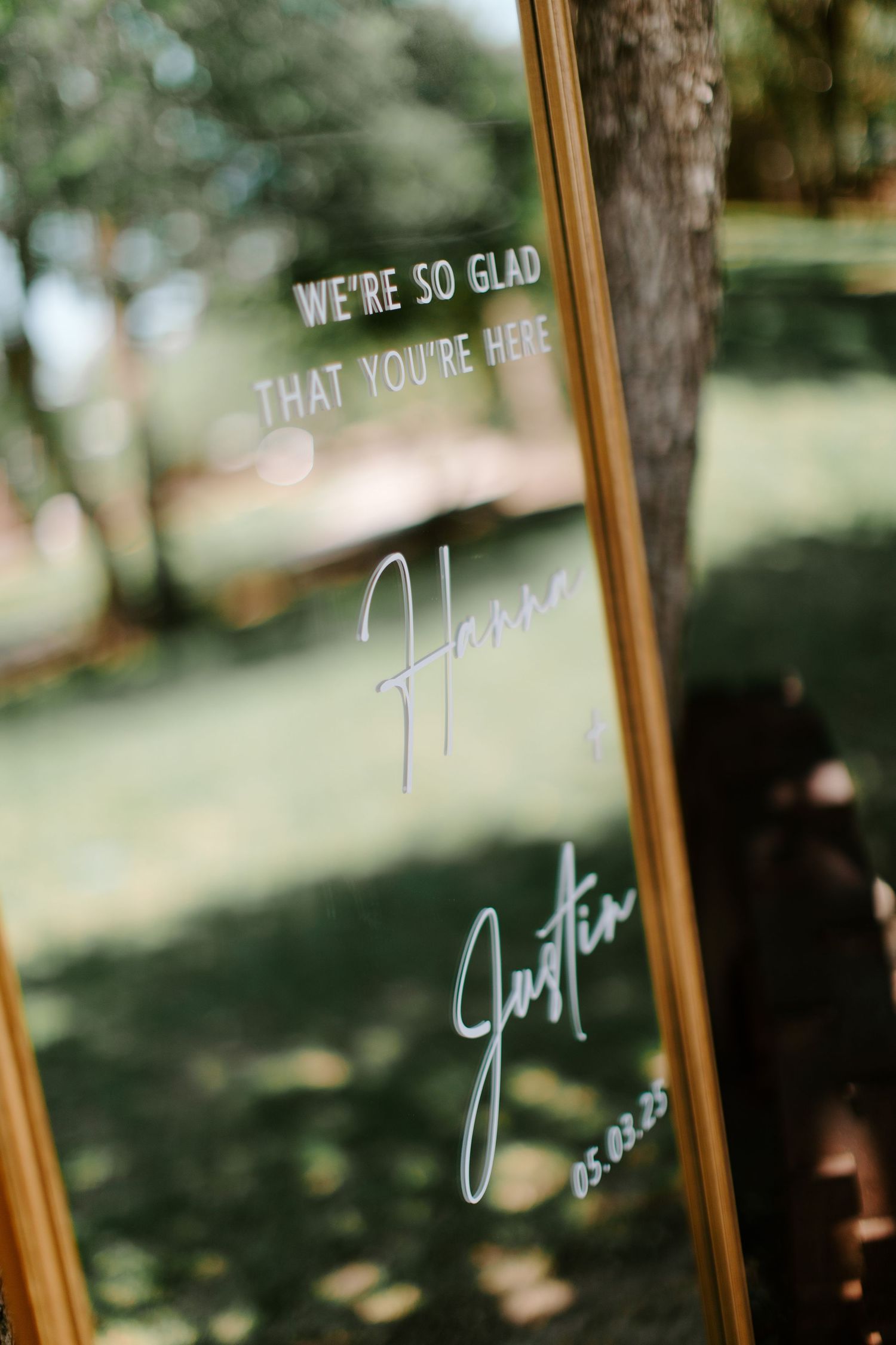 A welcoming glass sign with white text reading 'We're so glad that you're here' against a blurred natural background.