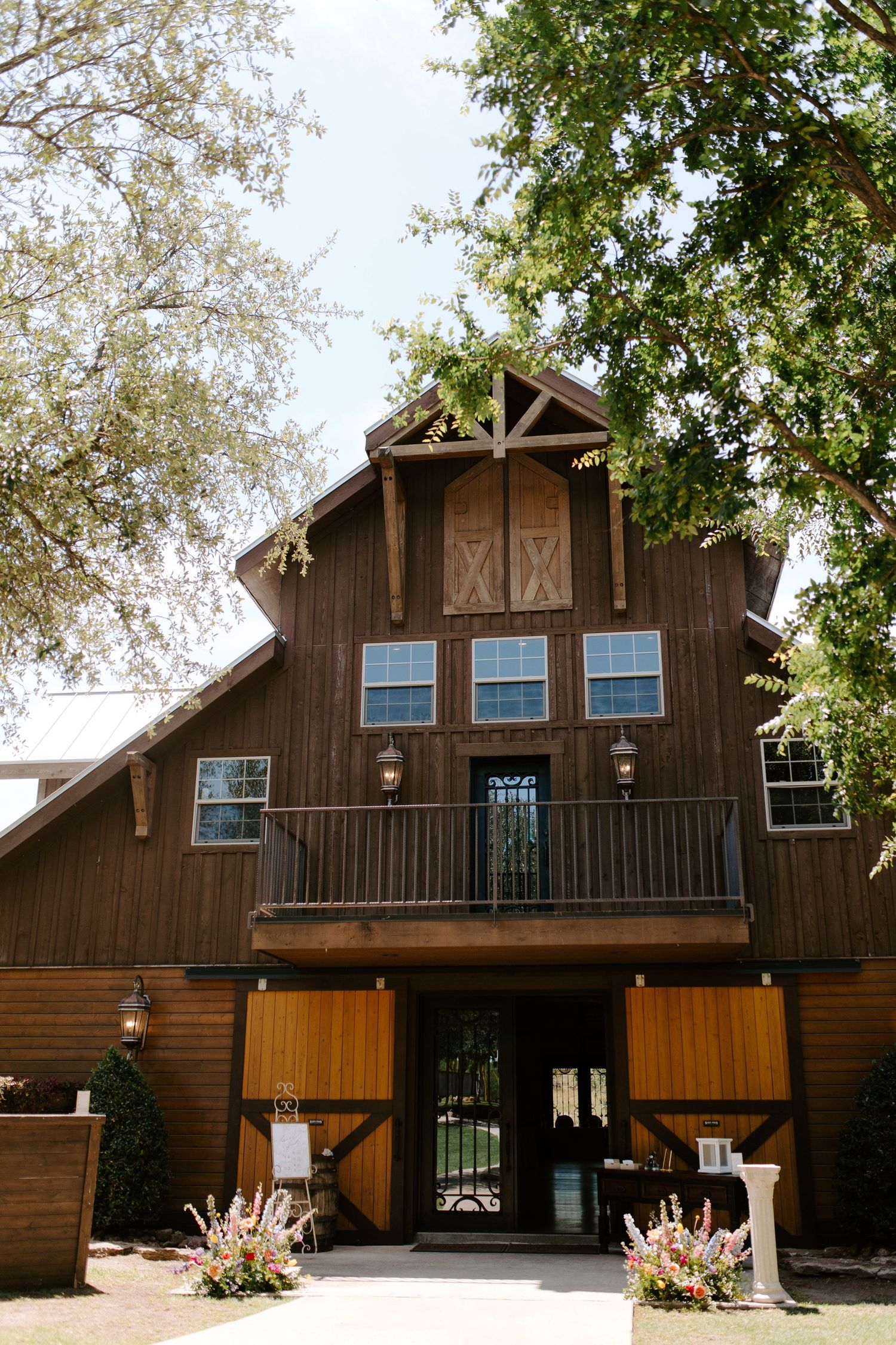 A rustic brown barn-style wedding venue with white trim and large wooden doors surrounded by trees.