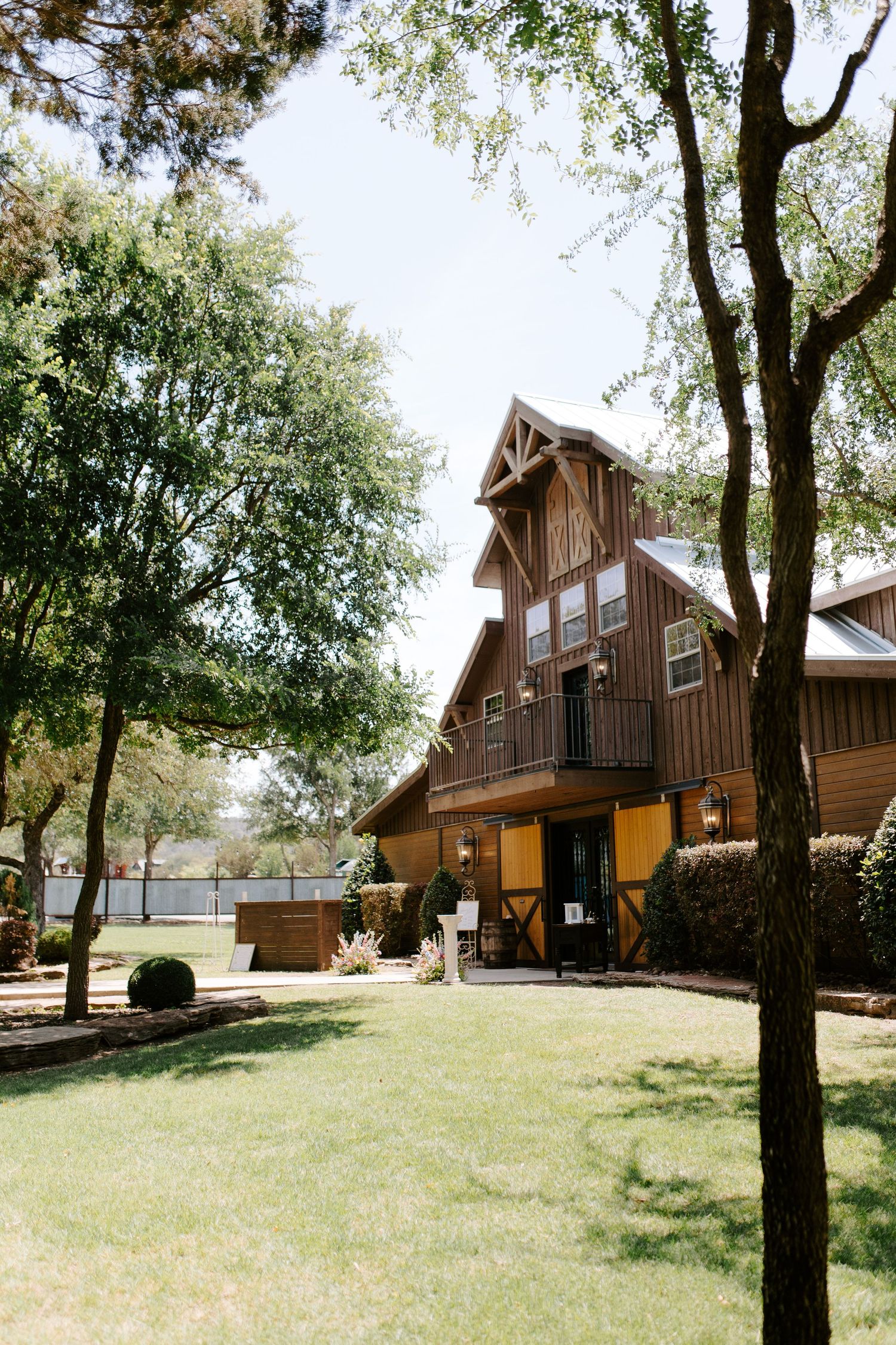 Rustic wooden barn-style wedding venue with tall trees and manicured lawn on a sunny day.