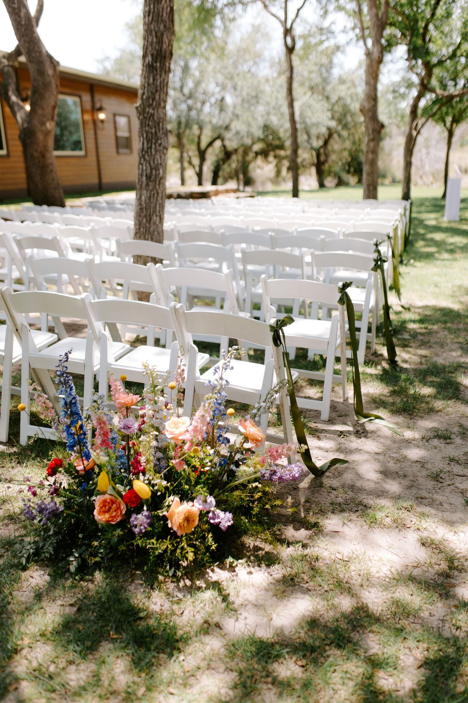 White wedding ceremony chairs arranged outdoors with colorful floral arrangements in dappled sunlight.