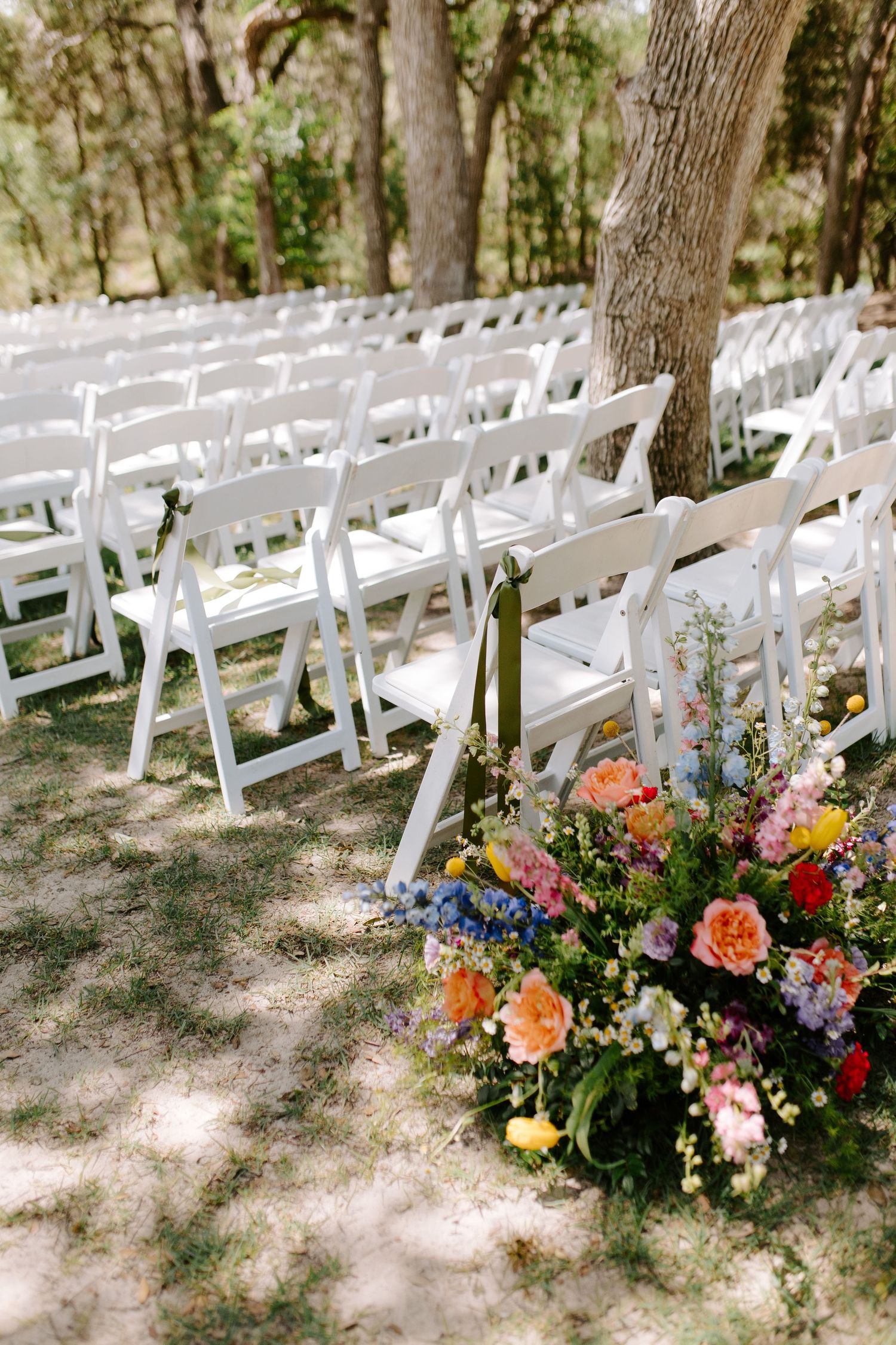 White folding chairs arranged for an outdoor wedding ceremony with colorful floral arrangements along the aisle.