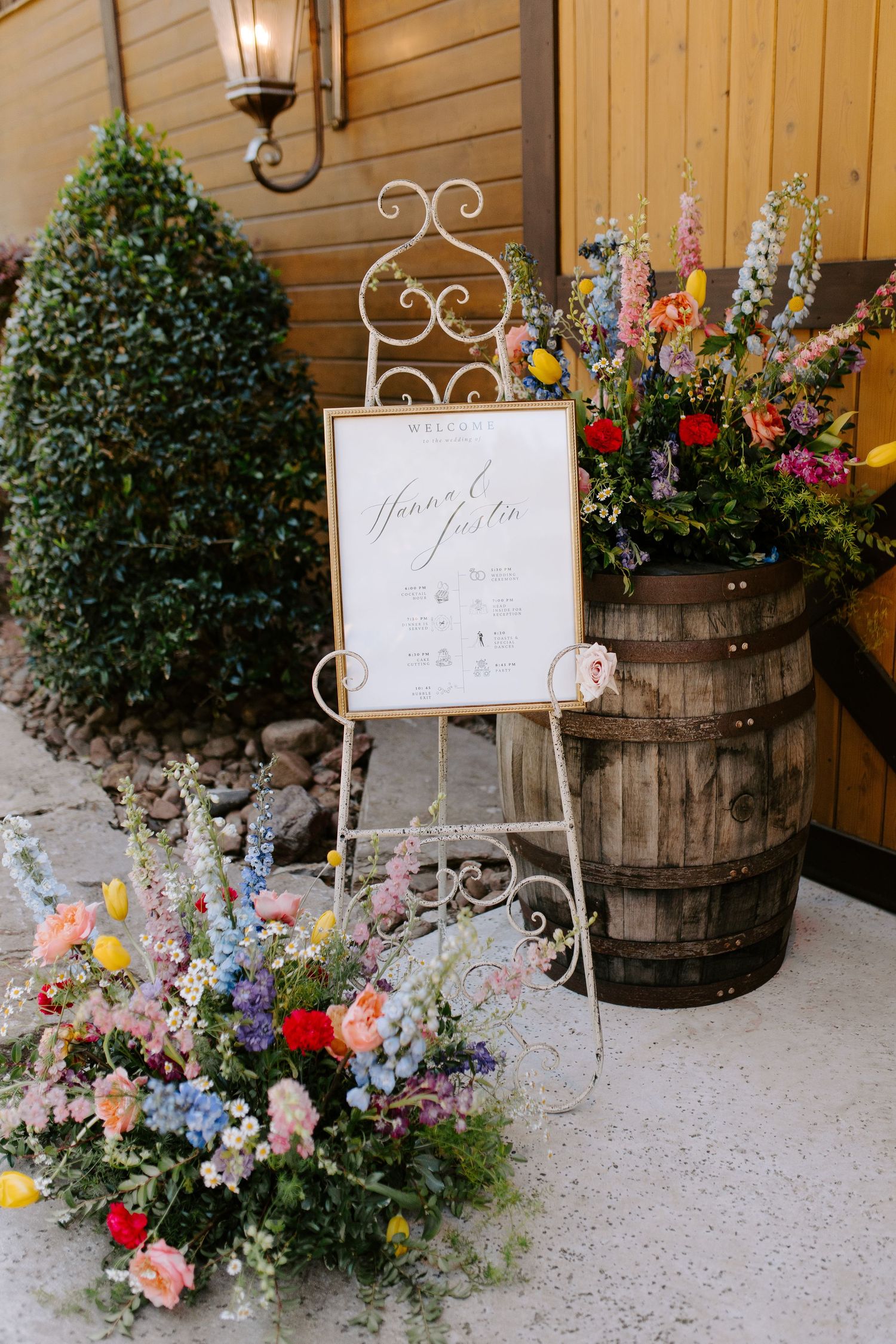 Rustic wedding decor featuring floral arrangements, wooden barrels and a welcome sign on an ornate easel.