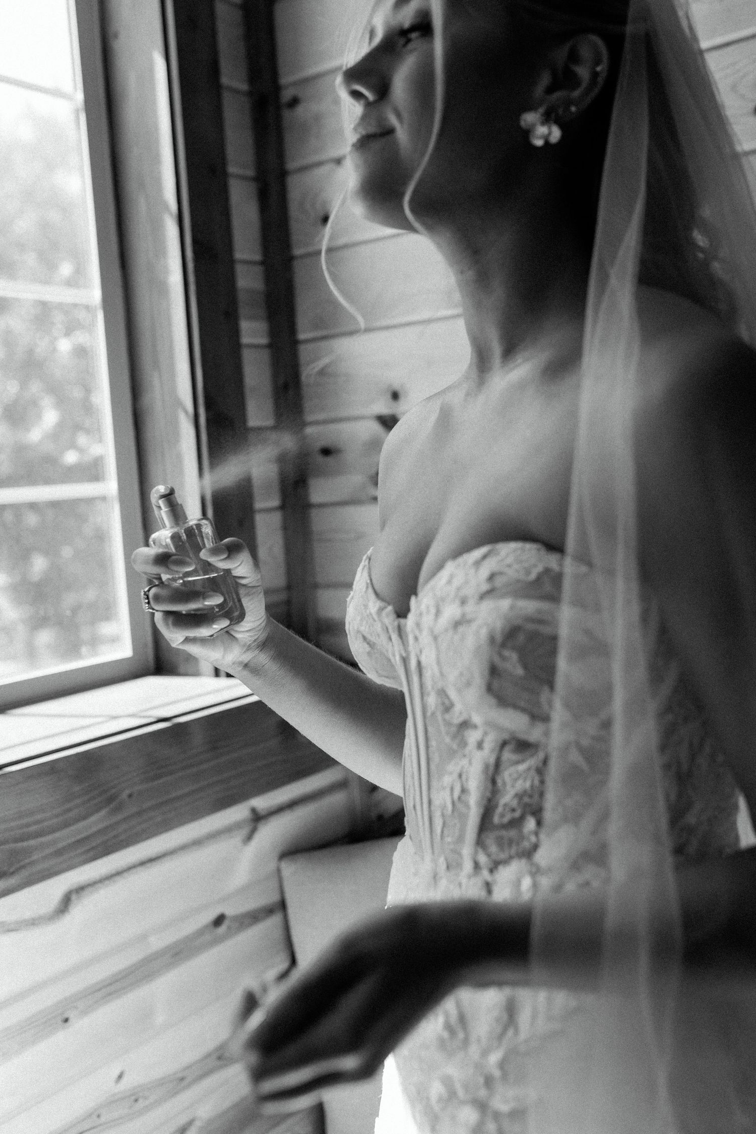 An artistic black and white photo of someone in a wedding dress by a window holding perfume.