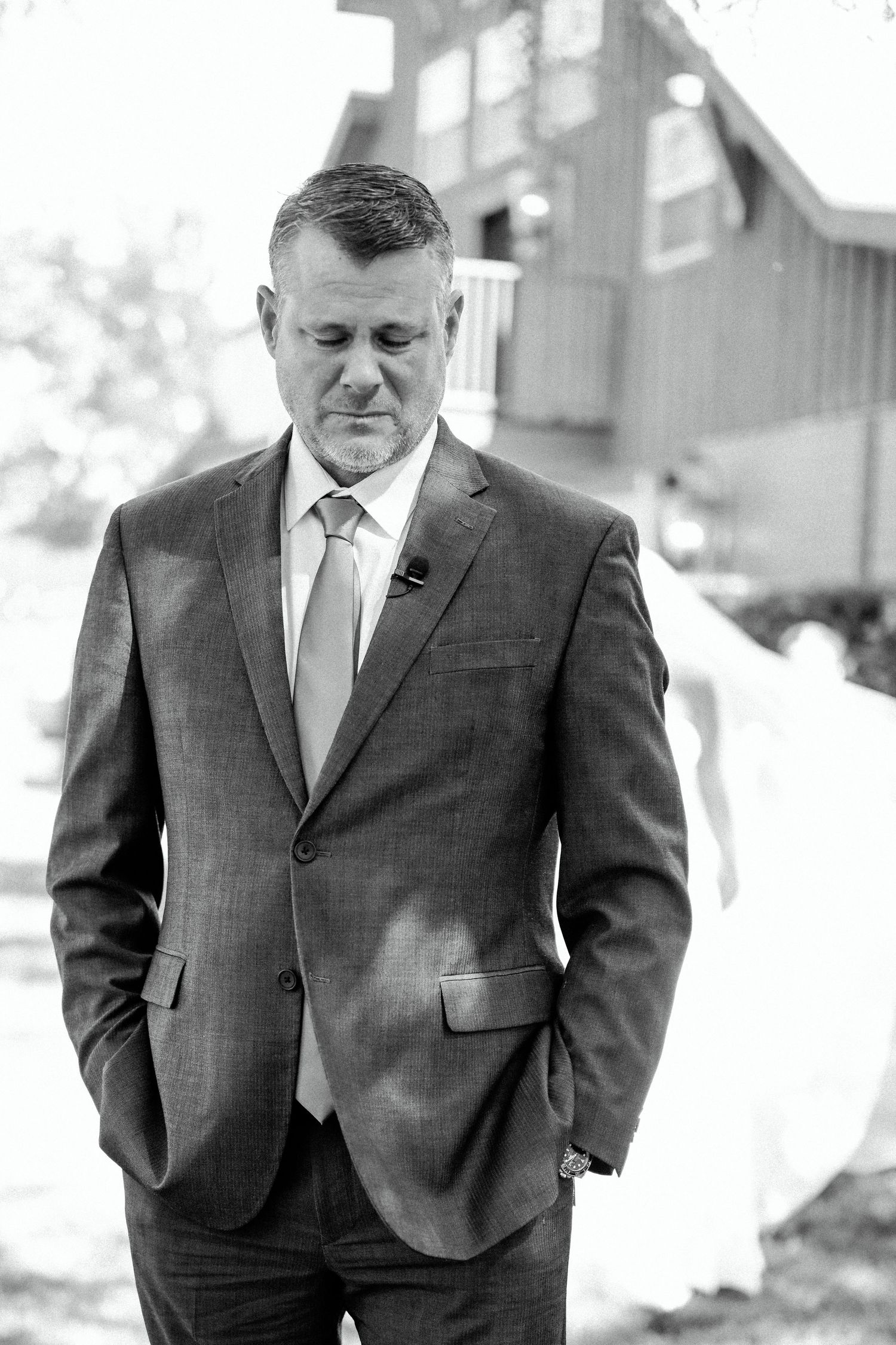 A person in a dark suit and tie stands with hands in pockets against a rustic outdoor backdrop.