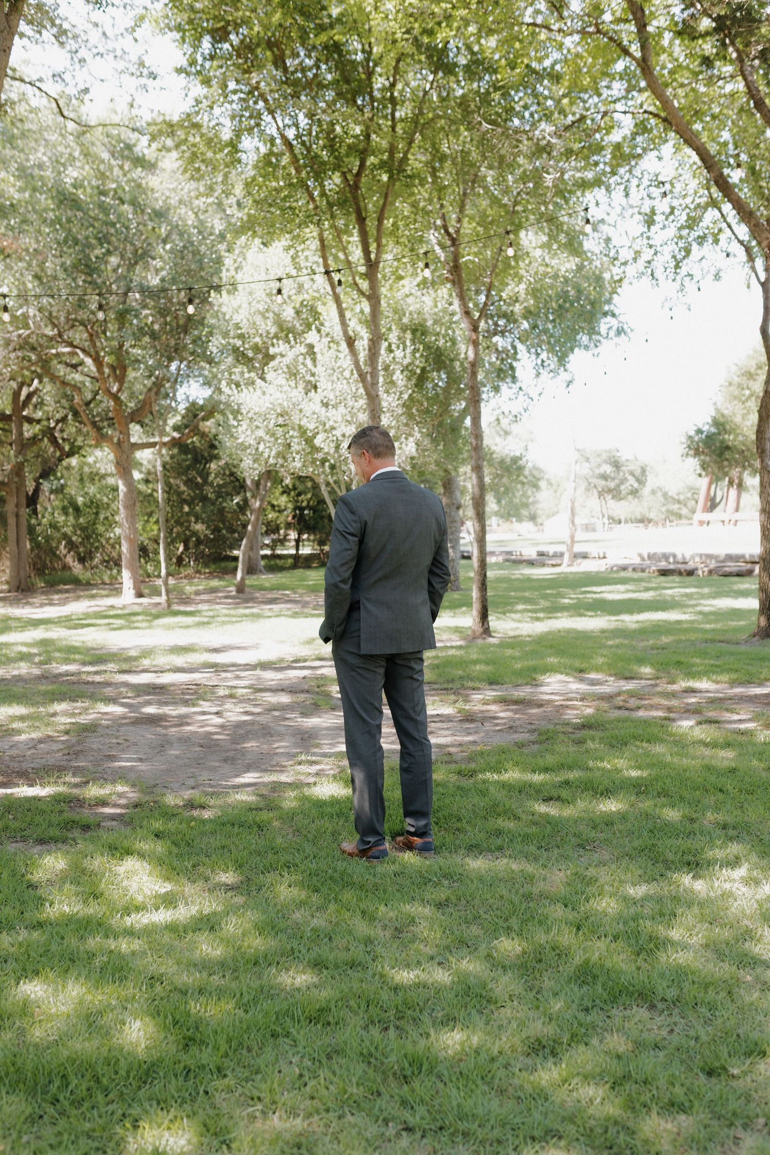A person in a dark suit standing alone on grass under shady trees.