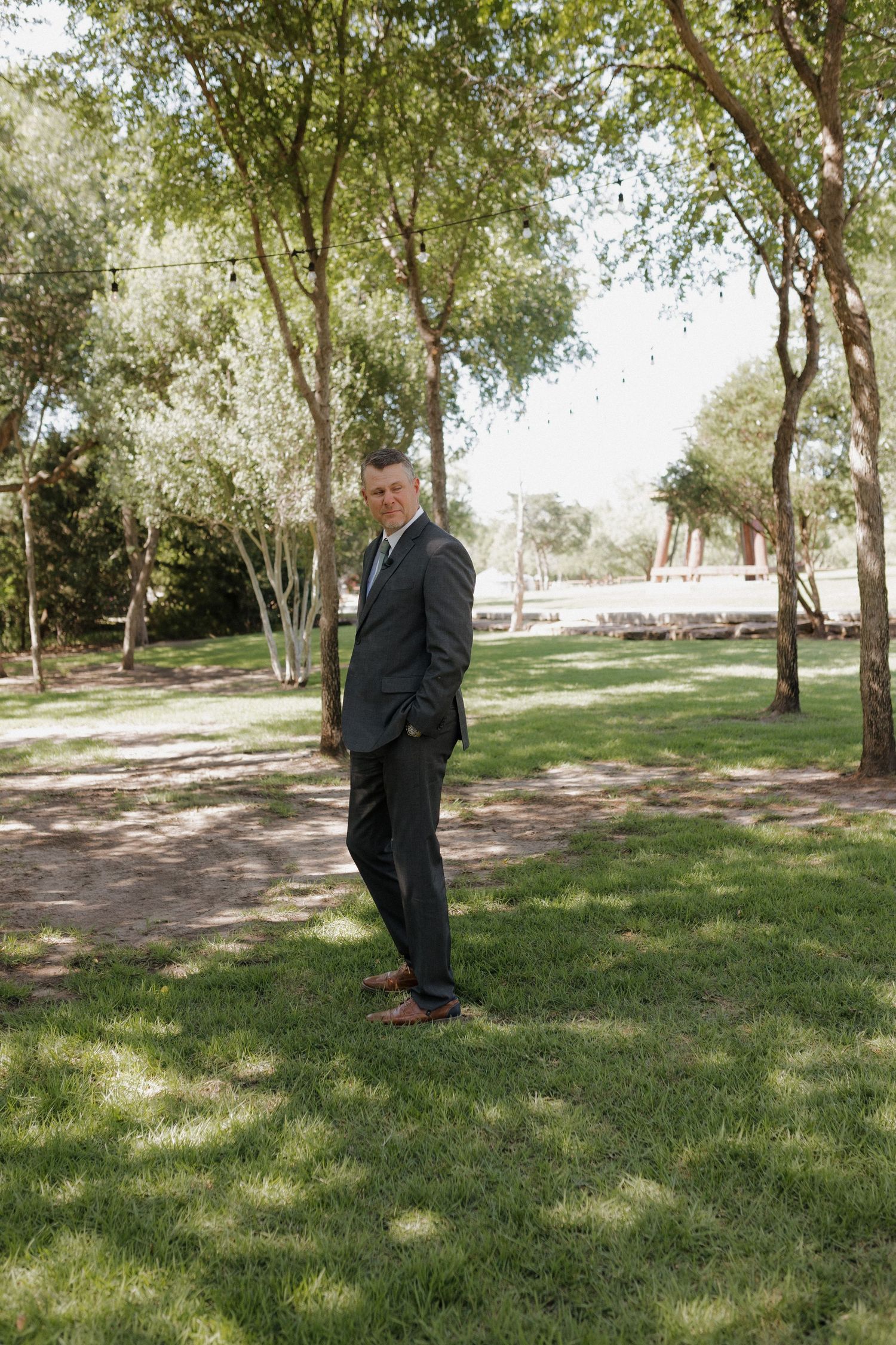 A person in a dark suit stands thoughtfully on green grass beneath tall trees in a peaceful park setting.