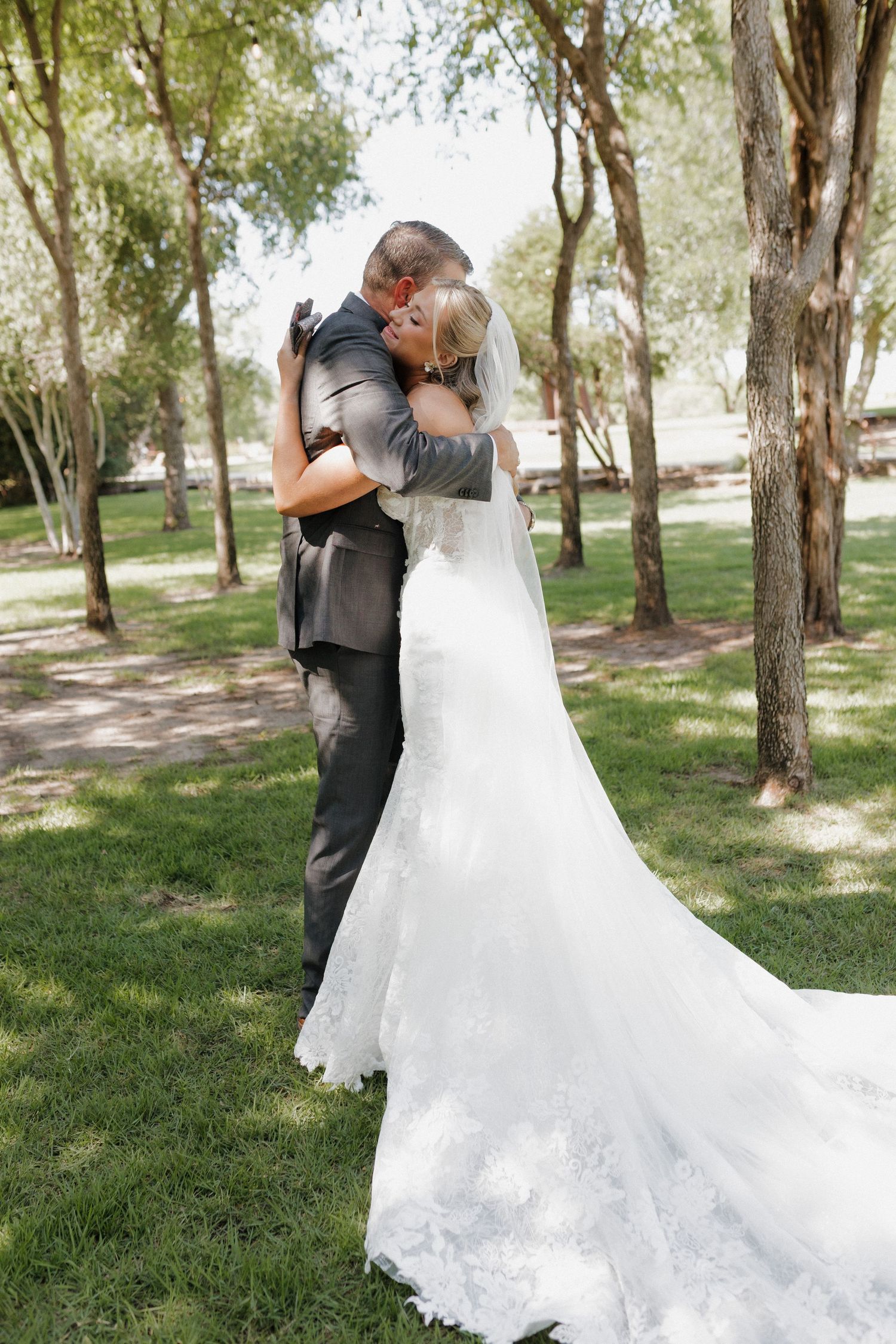 Wedding couple embracing outdoors among trees with the bride's white lace dress flowing in the background.