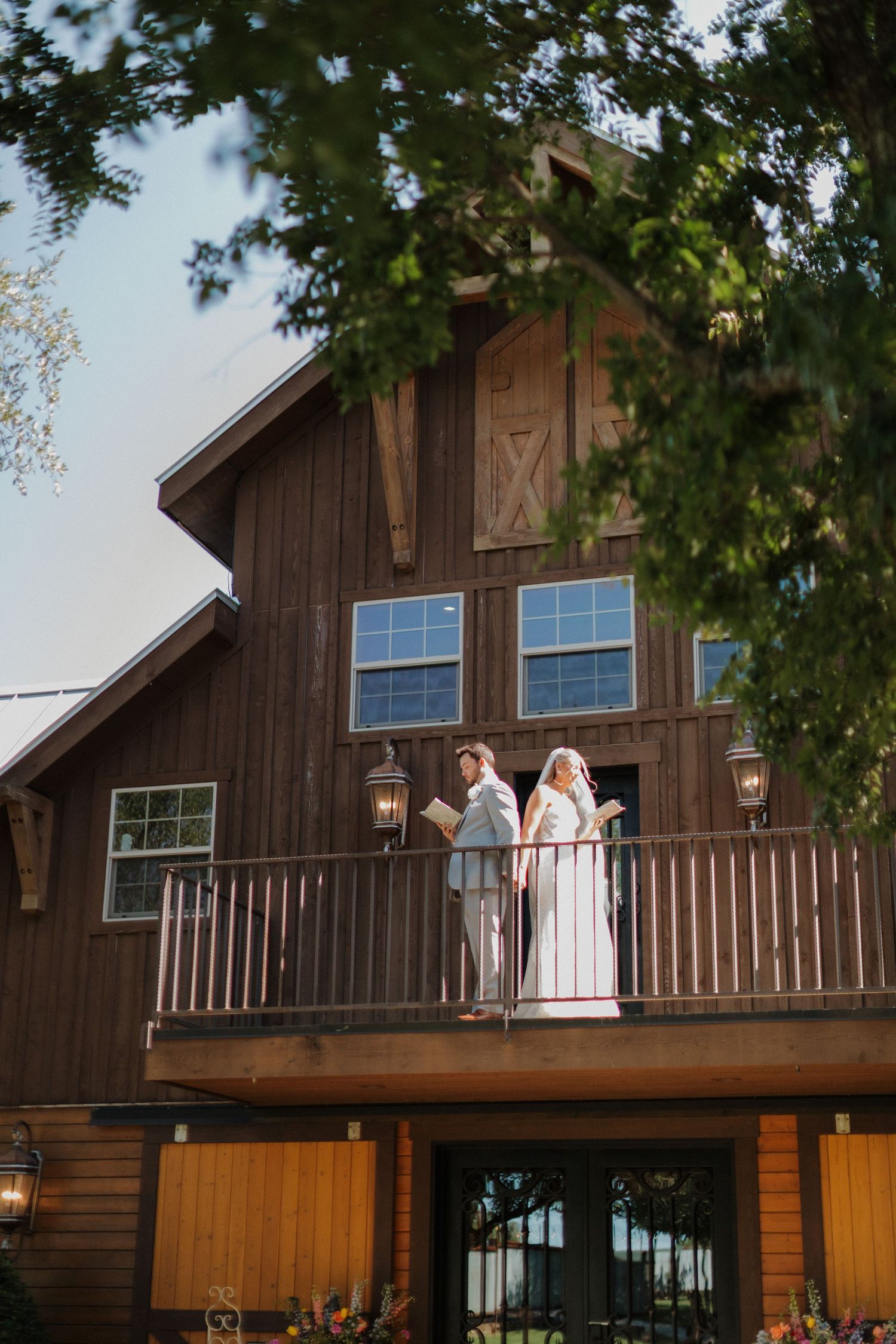Newlyweds stand on rustic barn balcony during golden hour at countryside wedding venue.