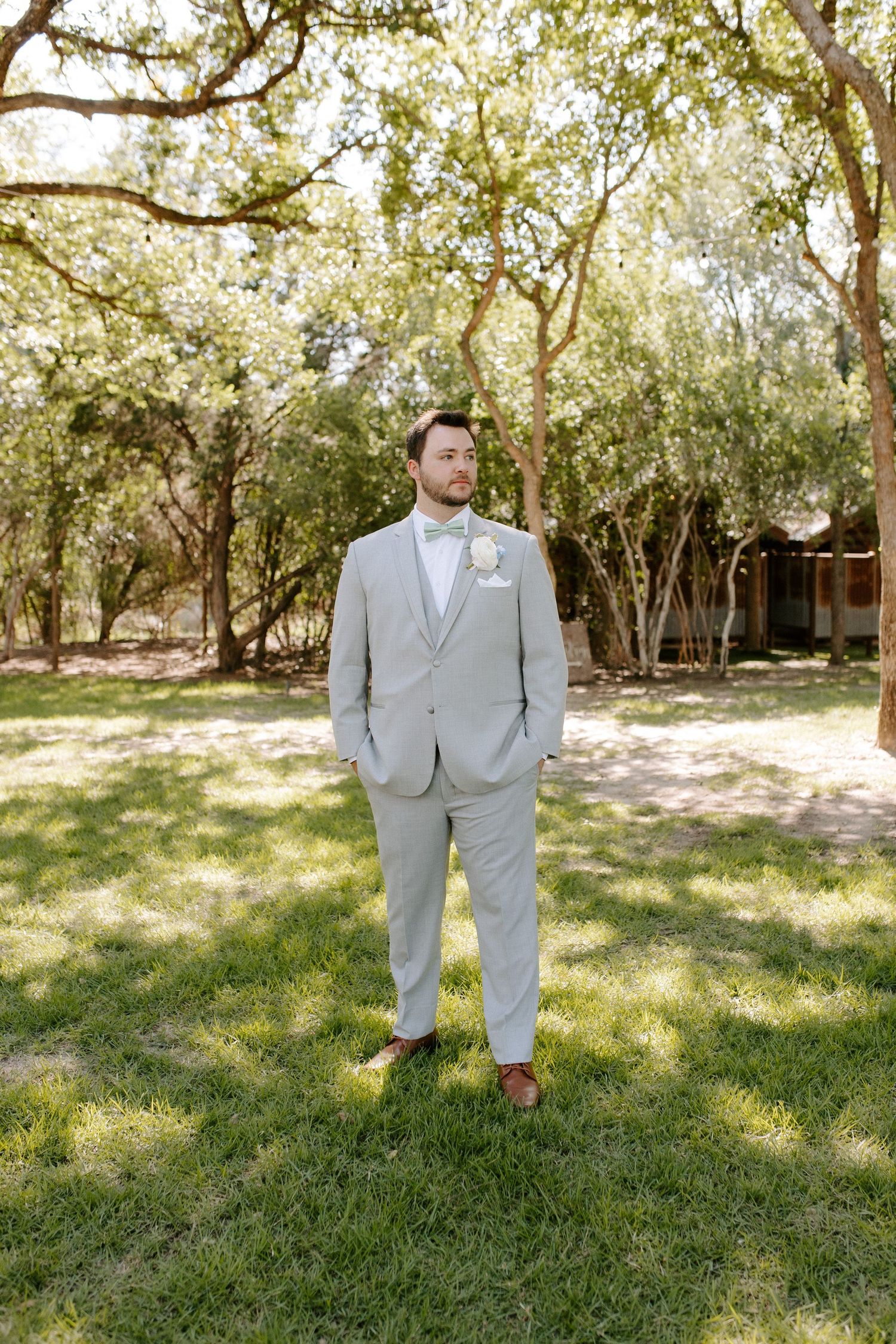 A groom in a light gray suit stands alone in a sunlit garden setting.
